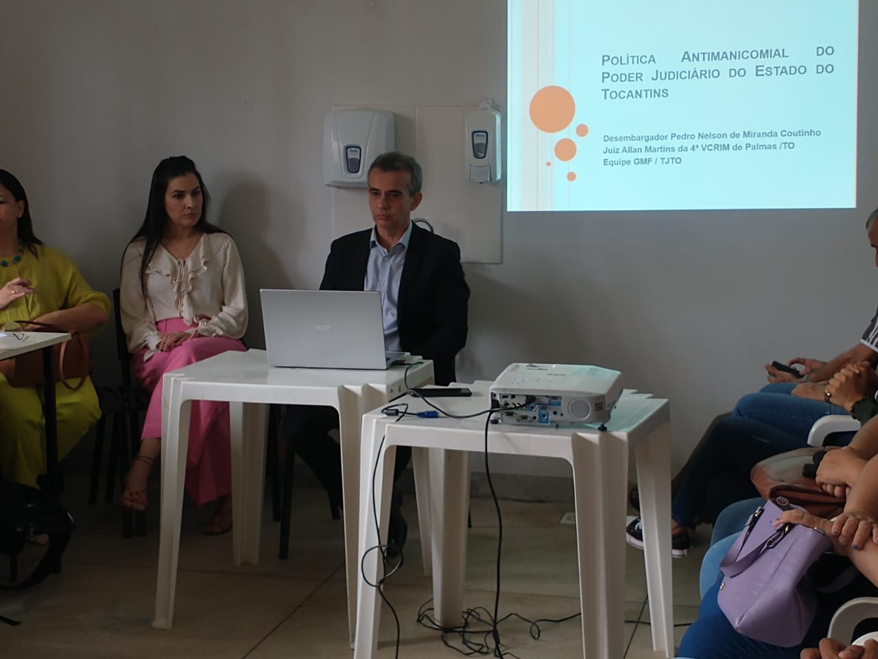 Juíz Allan Martins está sentado, na sua frente há um notebook e atrás um projetor refletindo uma imagem ampliada do que tem escrito no aparelho "Política Antimanicomial do Poder Judiciário do Estado do Tocantins".Há pessoas ao seu redor.