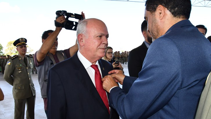 Homem calvo de terno e gravata, recebendo uma medalha de um homem moreno de terno