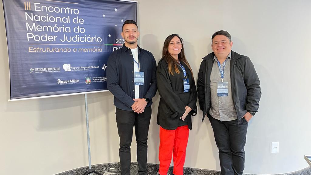 Imagem mostra três servidores, em pé, e atrás um banner sobre o Encontro Nacional da Memória do Poder Judiciário