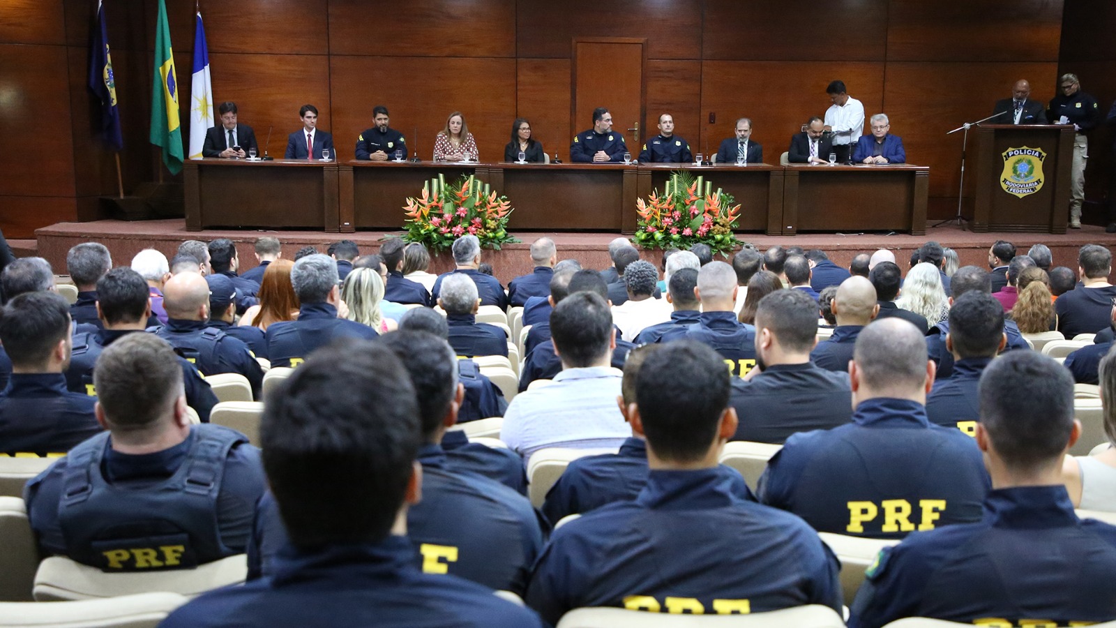Imagem mostra auditório lotado com pessoas sentadas e à frente uma mesa com autoridades prestigiando posse do superintendente da PRF