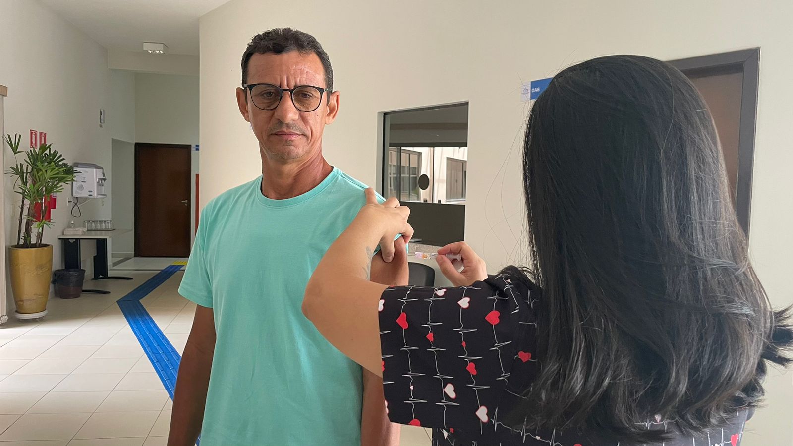 Mulher de cabelos lisos, de costas, aplica vacina em homem usa camisa verde e óculos de grau