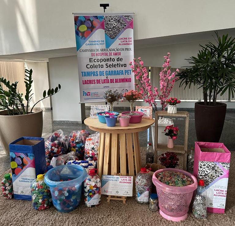 Tampinhas de várias cores e lacres de metal aparecem armazenados em caixas e baldes das cores azul e rosa, em um cenário de fundo branco