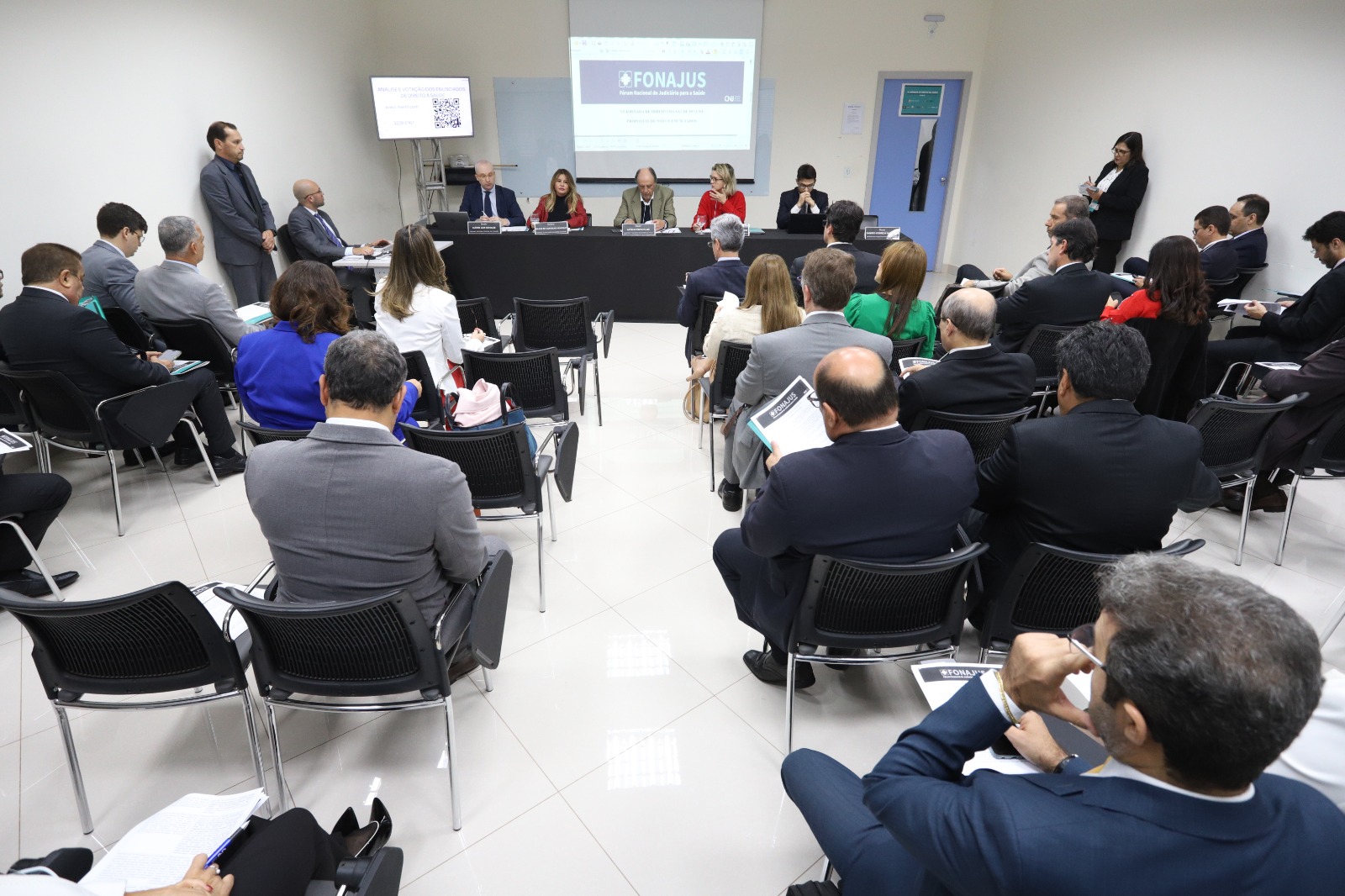 Imagem colorida que mostra pessoas sentadas em cadeiras numa sala fechada em frente a uma mesa