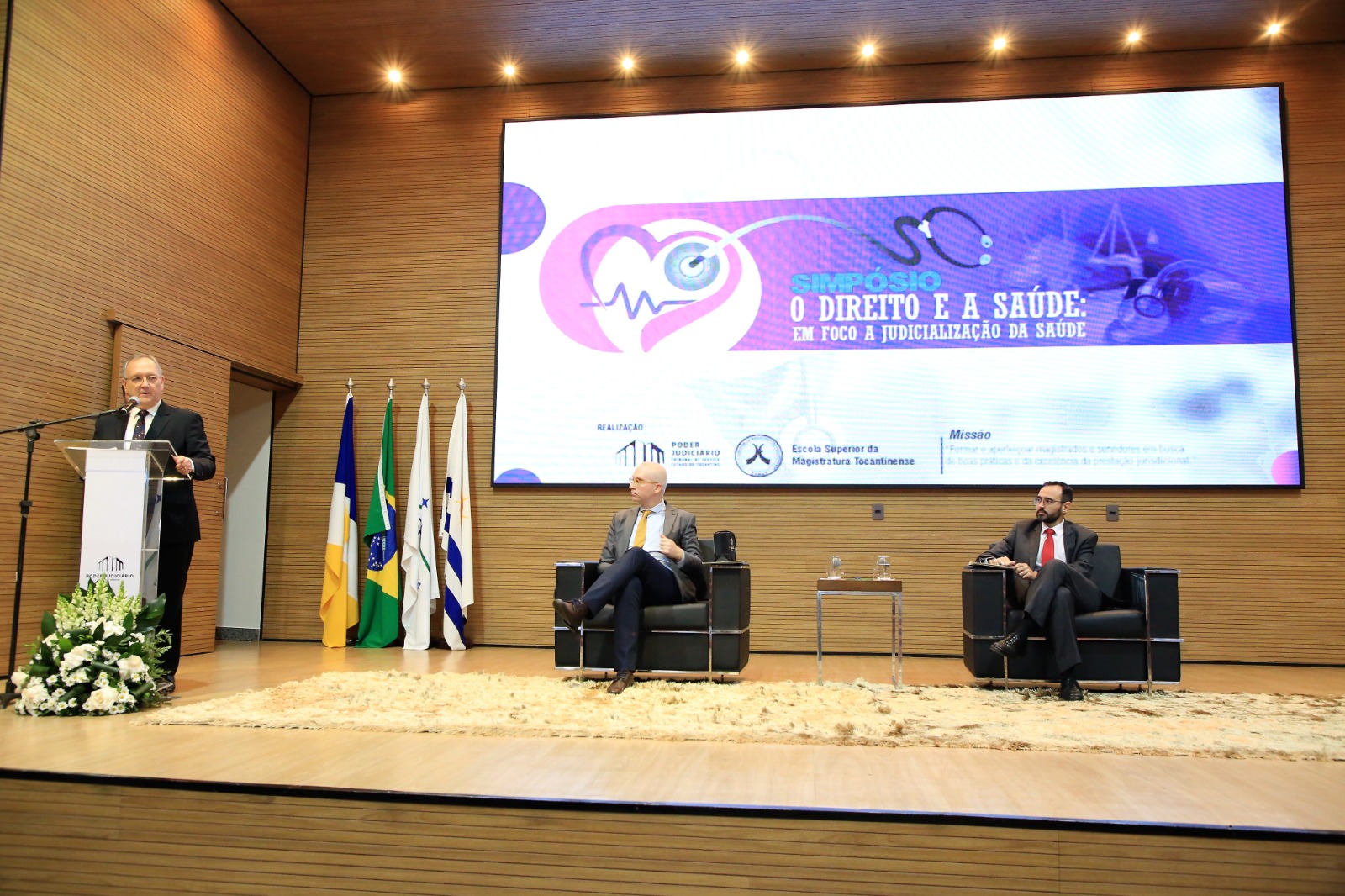 Em um palco, um homem branco, de terno,fala ao microfone enquanto é observado por outros dois homens de terno sentados em uma cadeira