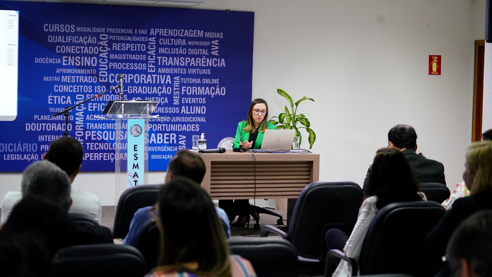 mulher de casaco verde fala ao microfone para plateia sentada