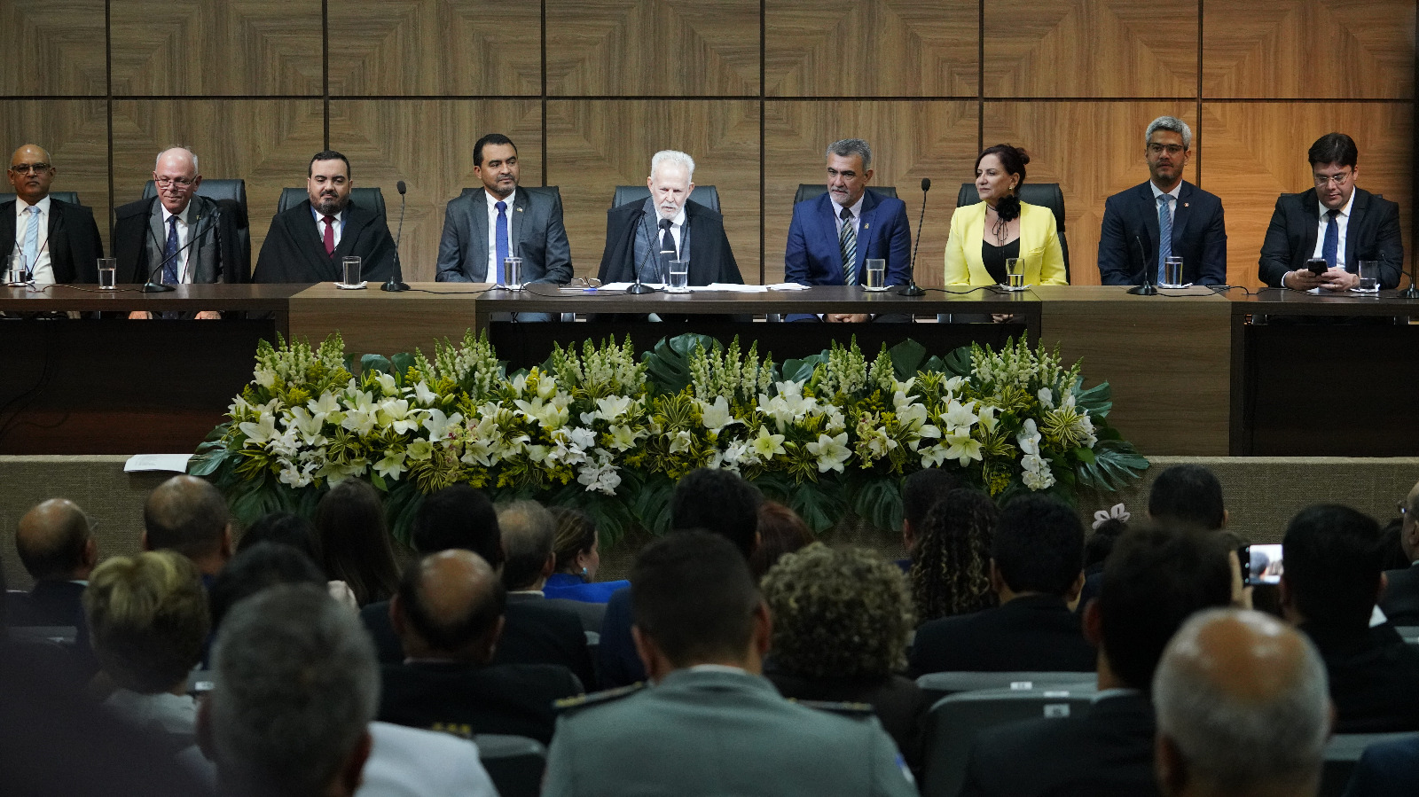 Numa mesa de autoridades encontram-se homens e mulheres bem trajados sentados atrás de um arranjo de flores e observados por uma platéia
