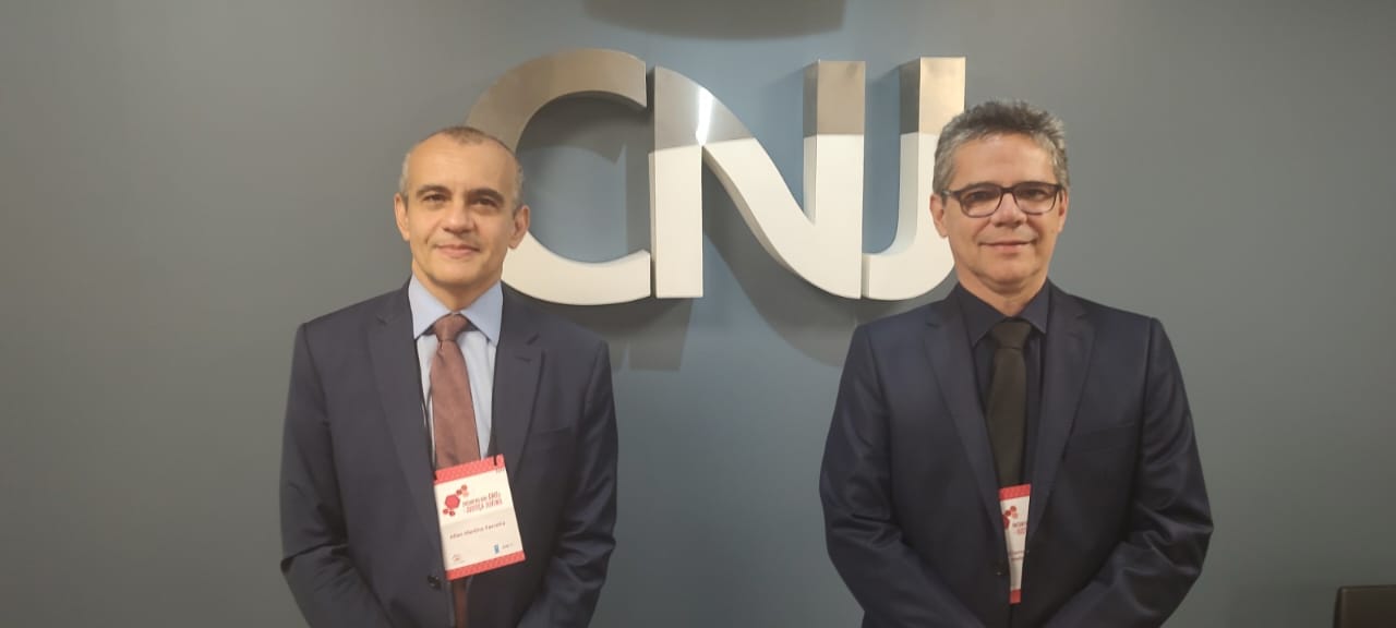 Foto mostra dois homens vestidos de terno escuro. Ao fundo parede cinza, escrito CNJ