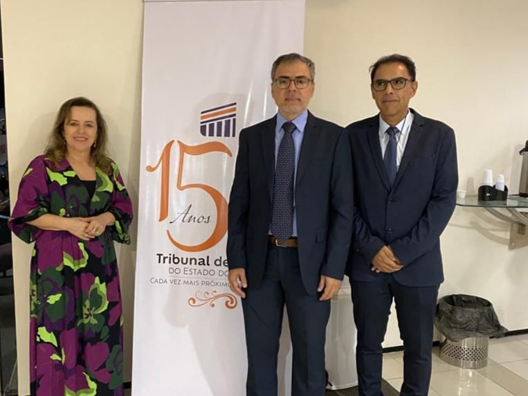 Uma mulher e dois homens posam para foto em pé ao lado de um banner durante Jornada Maria da Penha