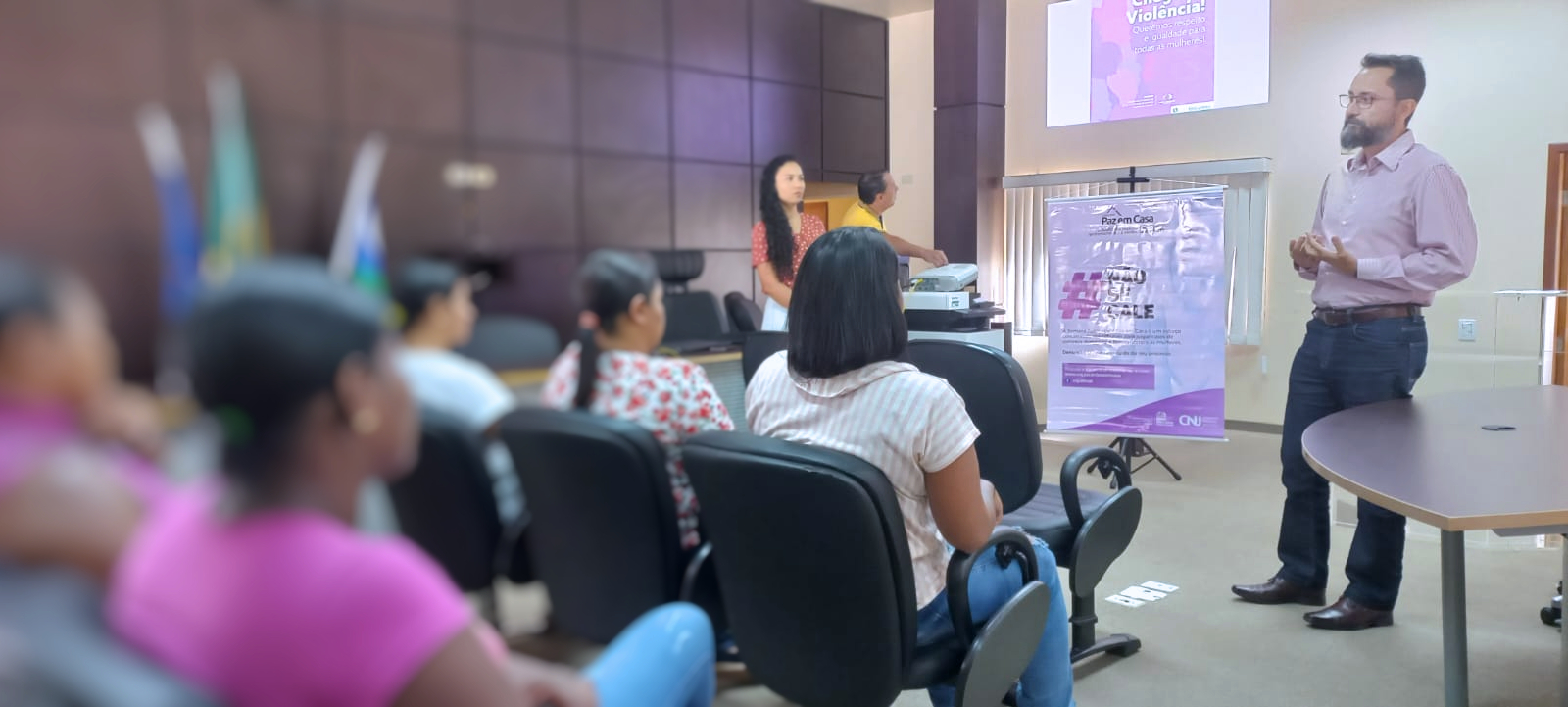 Imagem colorida mostra um homem jovem de barba falando para uma plateia de mulheres que não são identificadas 