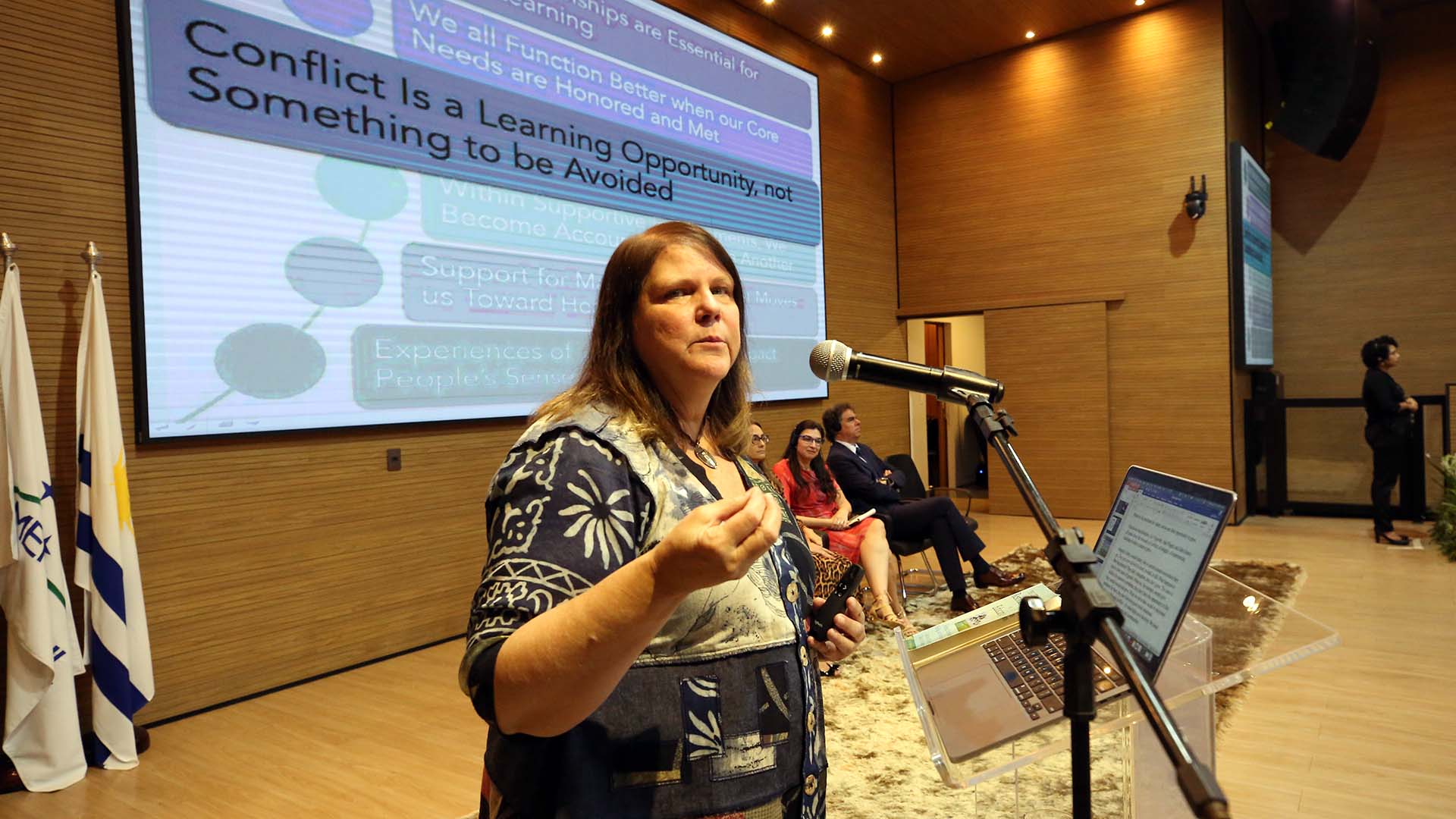 Mulher branca fala num púlpito  com imagens em um telão de fundo. 