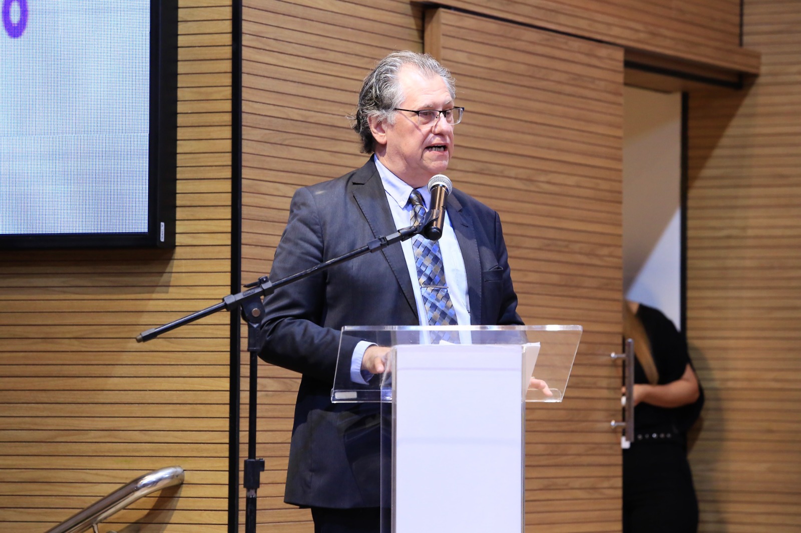 Imagem traz um homem alto, branco usando terno escuro atrás do púlpito transparente