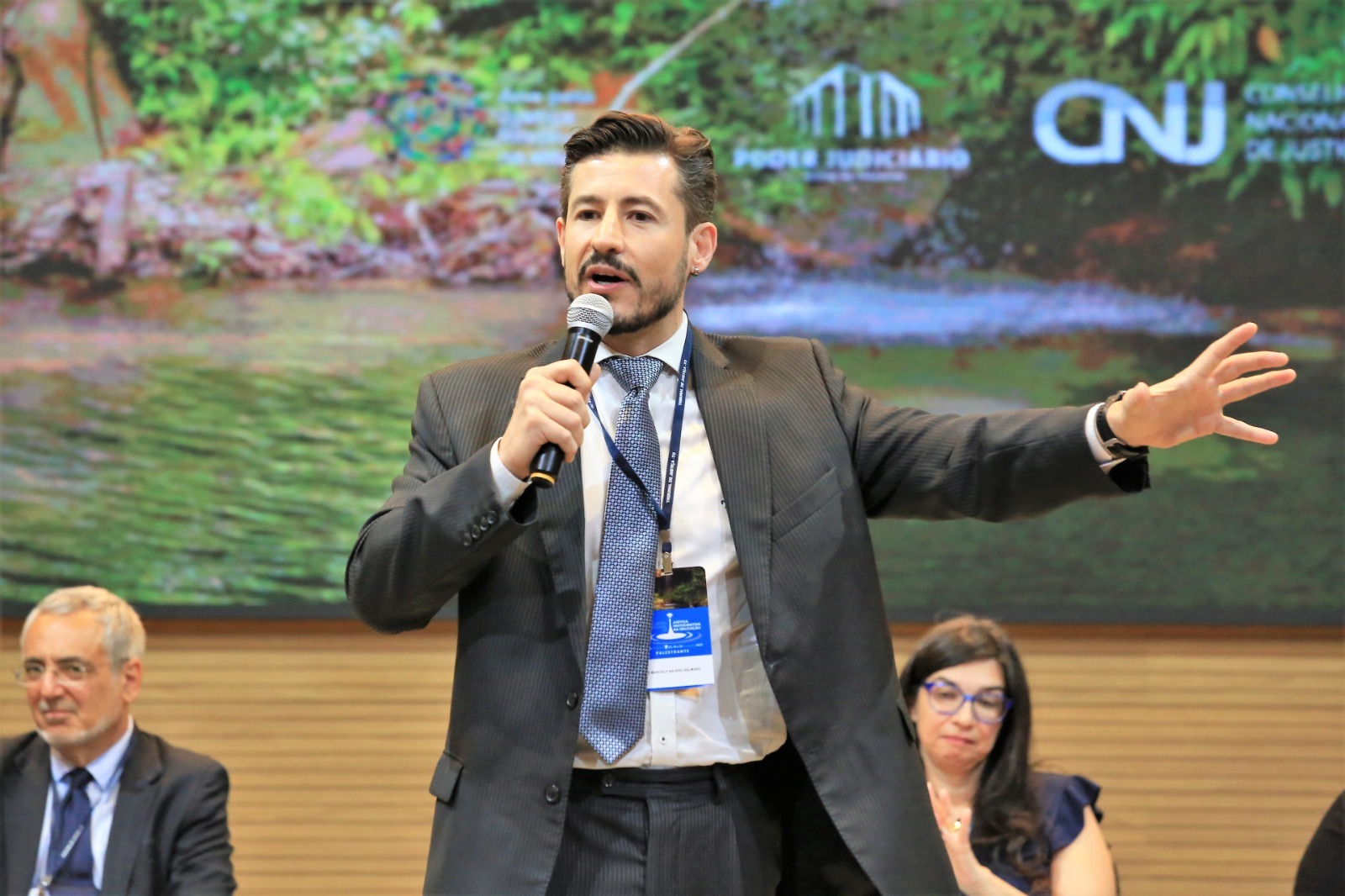Imagem mostra um homem branco, usando terno e segurando um microfone durante palestra