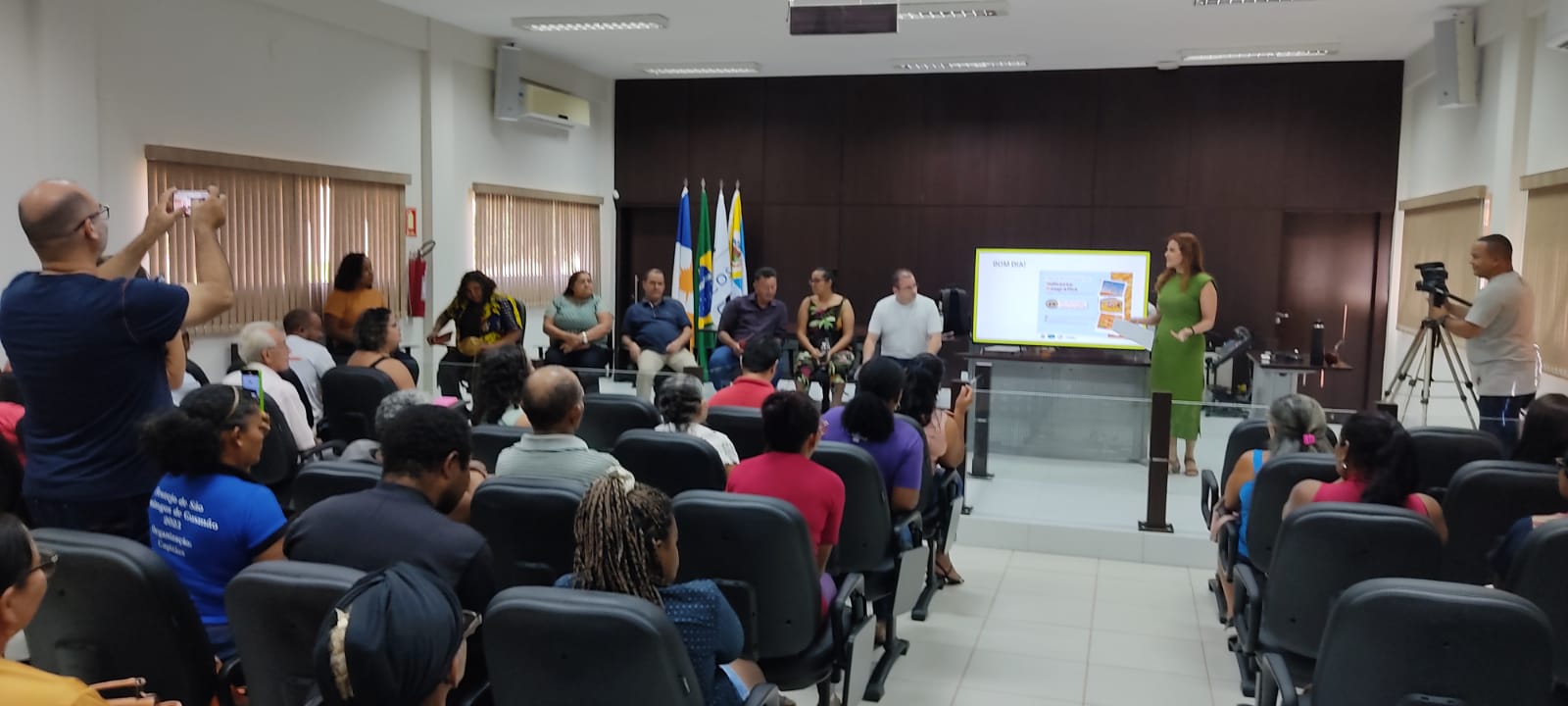 Juíza de vestido verde, em pé na frente de uma tv, no auditório ministrando oficina para homens e mulheres sentados