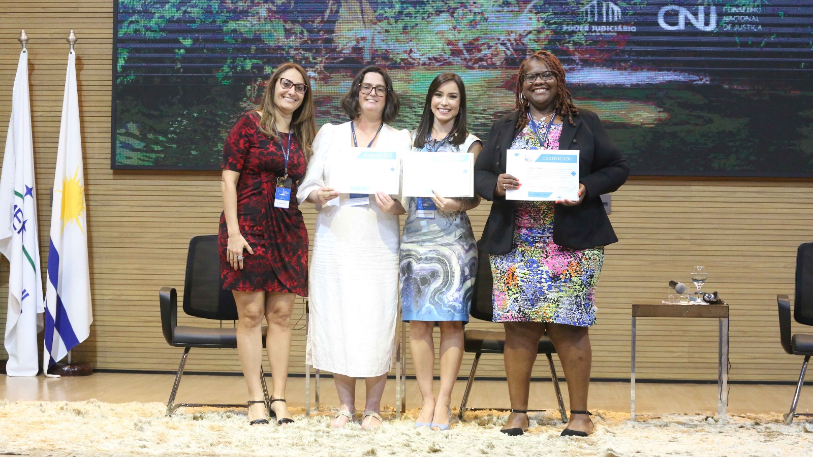 Mulheres em pé recebendo certificados, ao fundo quadro com foto de cachoeira e bandeiras na lateral