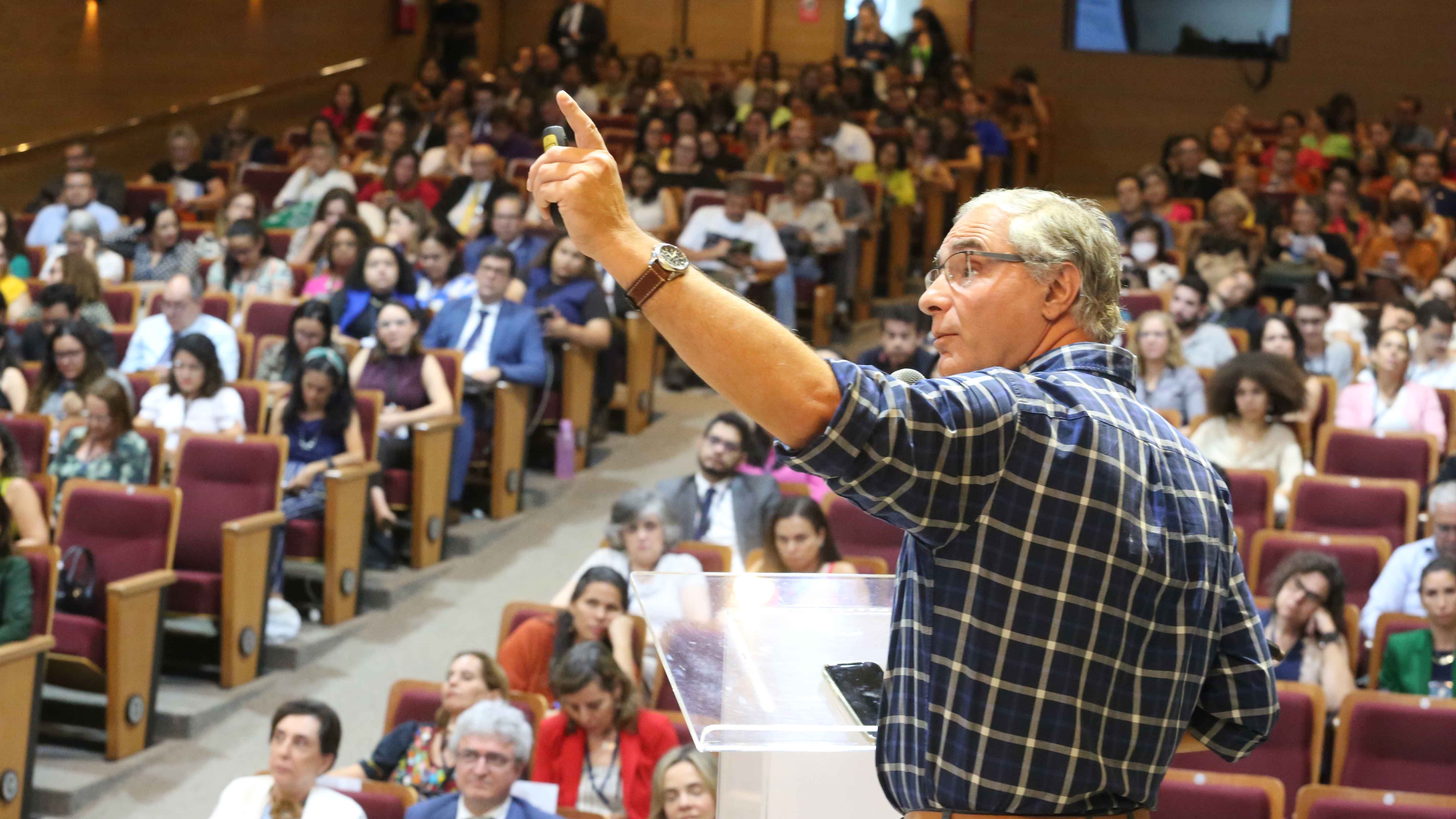 Palestrante de camisa de xadrez azul em primeiro plano, ao fundo auditório cheio de pessoas sentadas