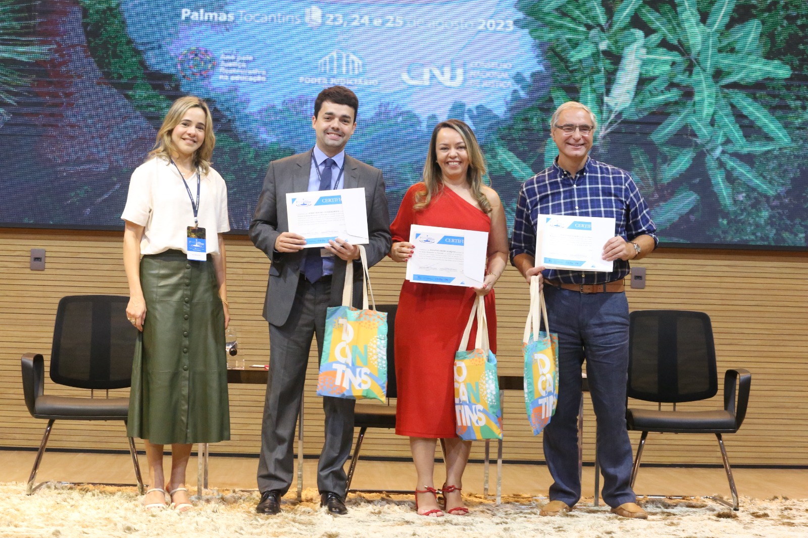 Homens e mulheres em pé segurando certificados e bolsas em cima do palco em encerramento de palestra