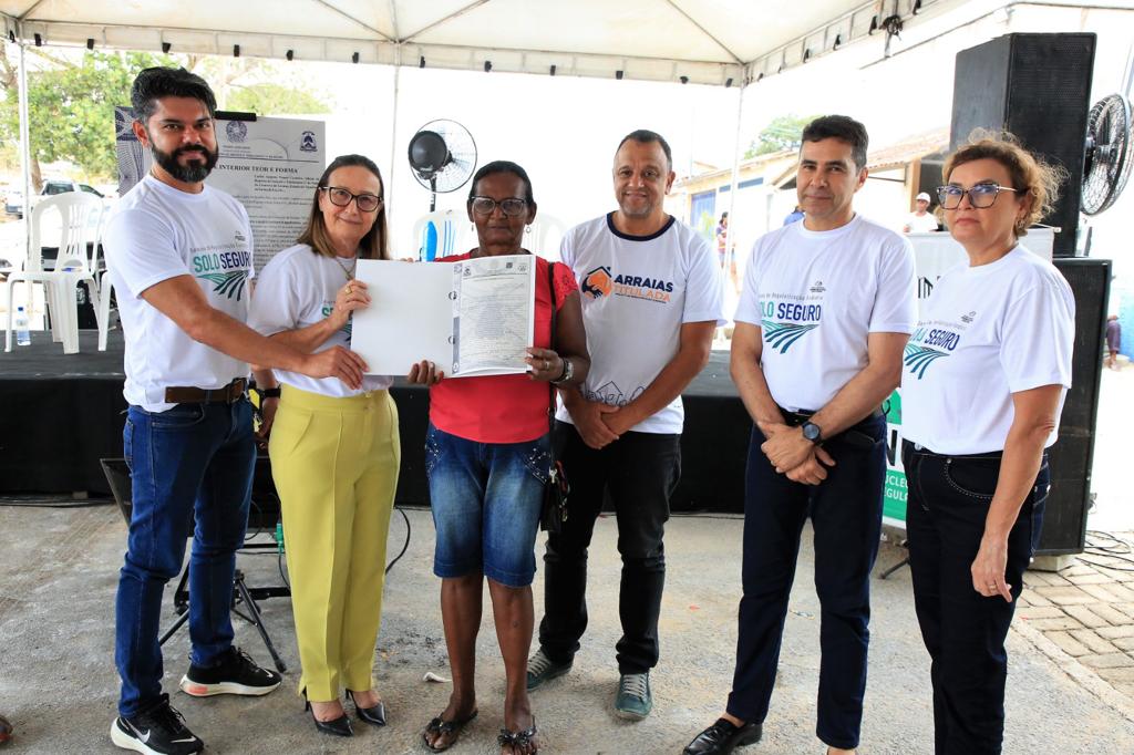 Imagem com cinco pessoas em pé, e duas mulheres segurando um papel de um título de propriedade