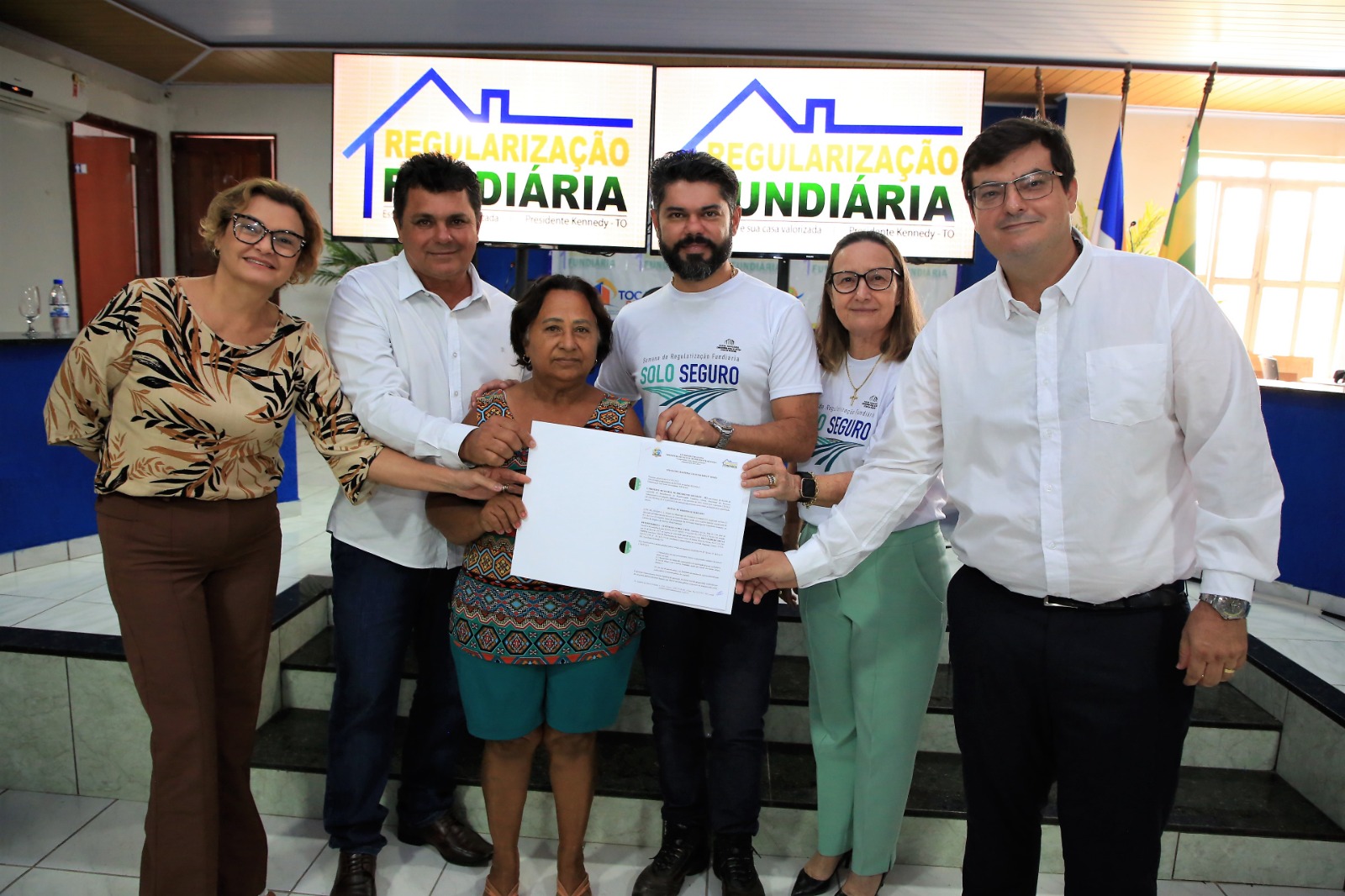 Autoridades segurando título de propriedade juntamente a mulher beneficiada e posando para foto