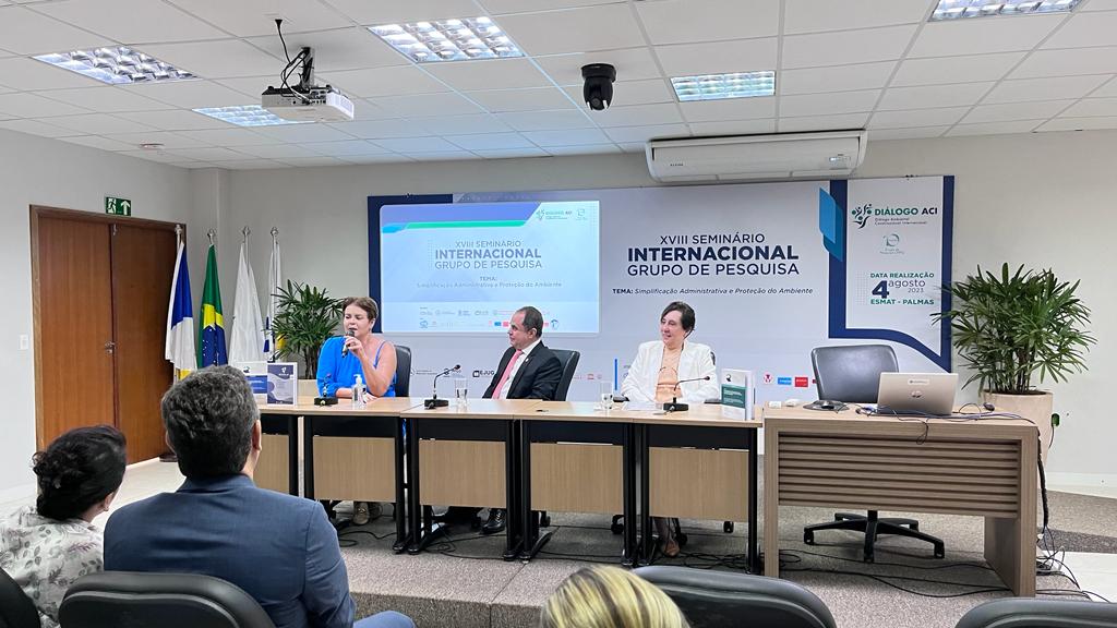 Fotografia colorida de autoridades sentados à mesa enquanto são assistidos pela plateia 