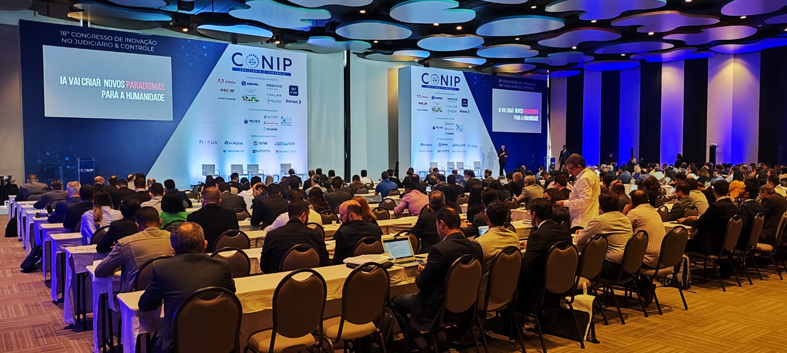 Homens e mulheres sentadas em ambiente fechado assistindo palestra sobre tecnologia