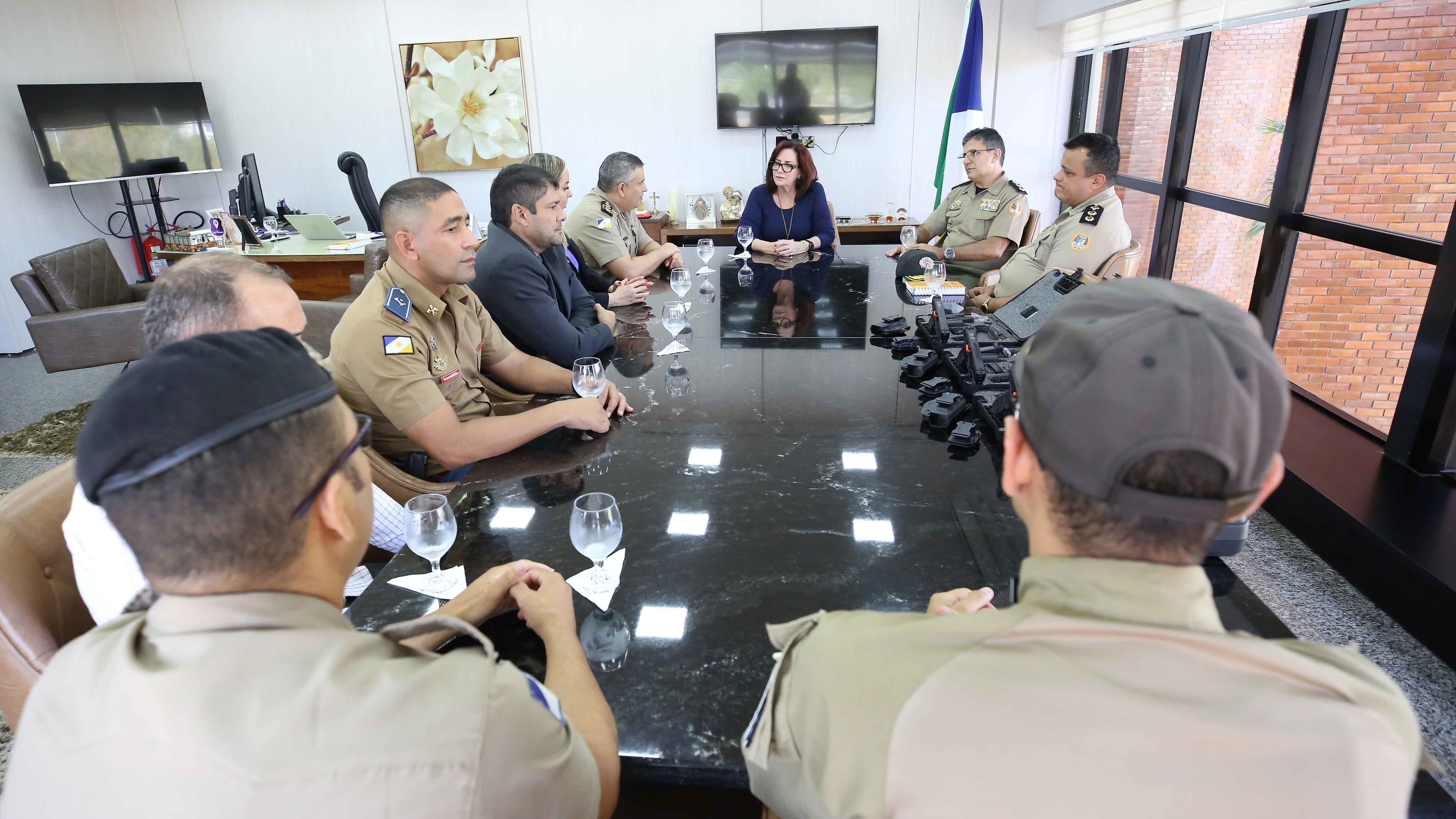 Pessoas sentadas ao redor de uma mesa, na sala da presidência, e em cima da mesa existem copos de vidro e as armas a serem entregues