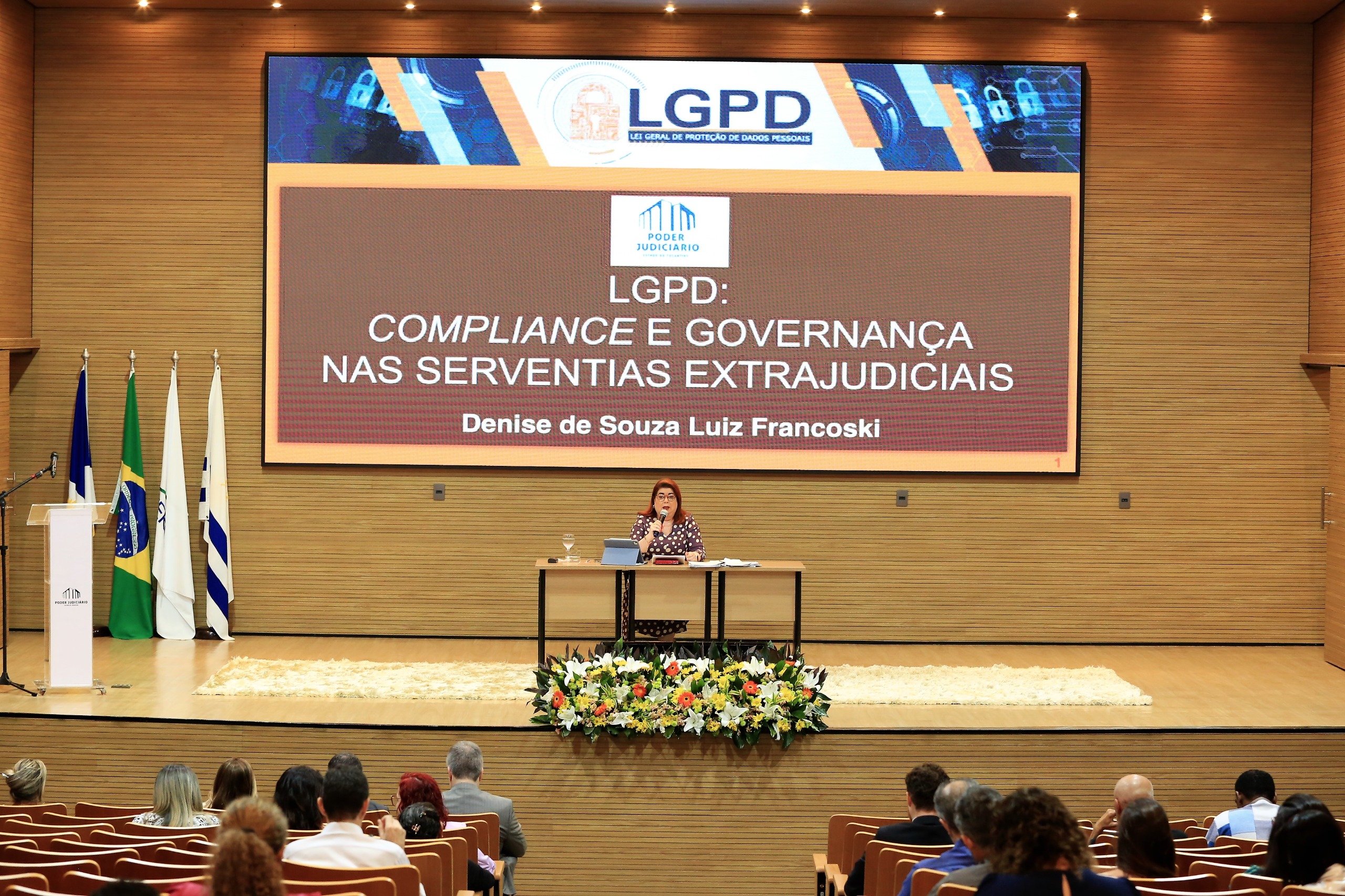 mulher sentada em mesa falando ao microfone para o público com slide projetado as suas costas 