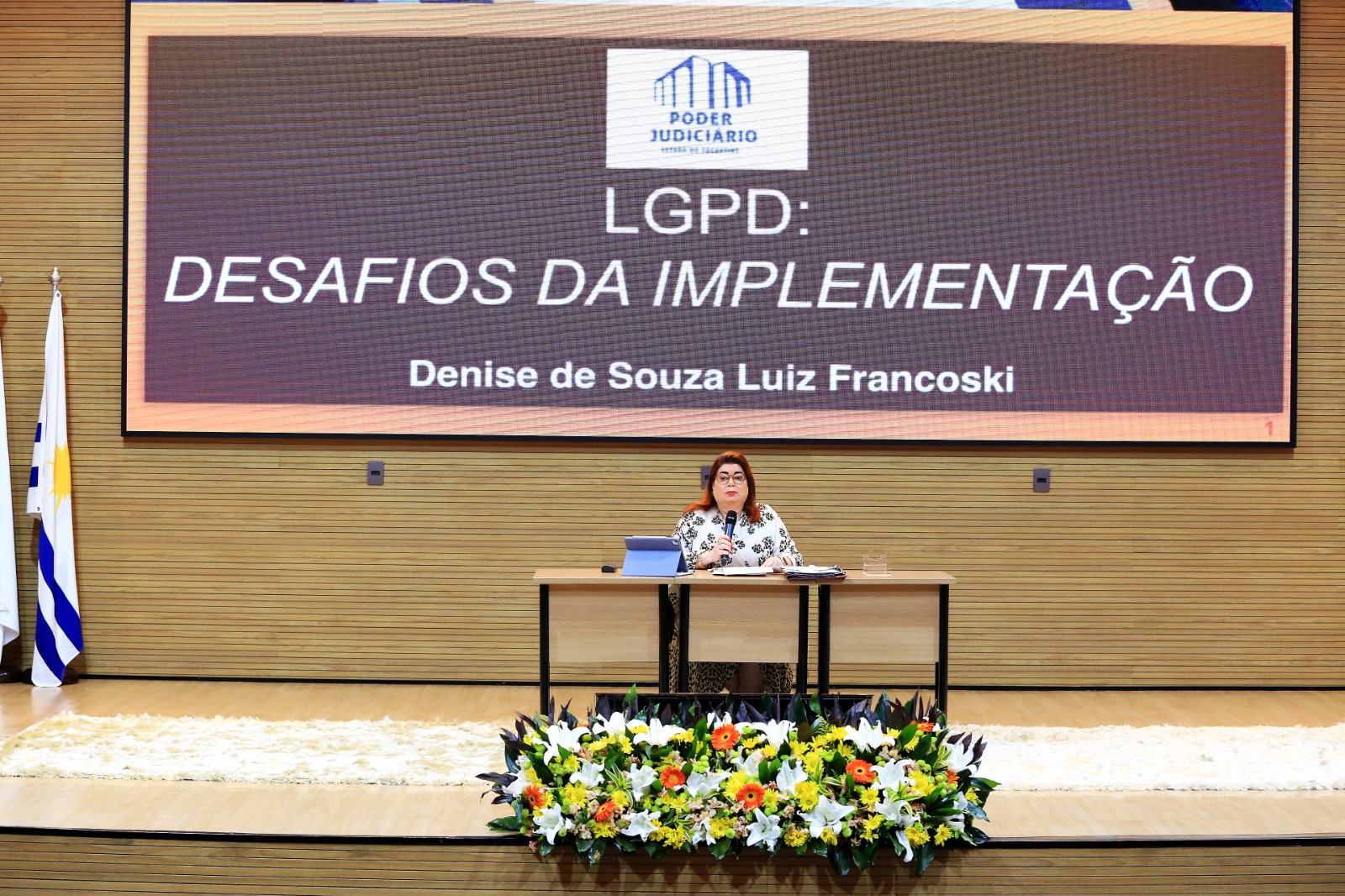 mulher sentada e falando ao microfone com slide projetado atrás dela com as escritas "LGPD: DESAFIOS DA IMPLEMENTAÇÃO"