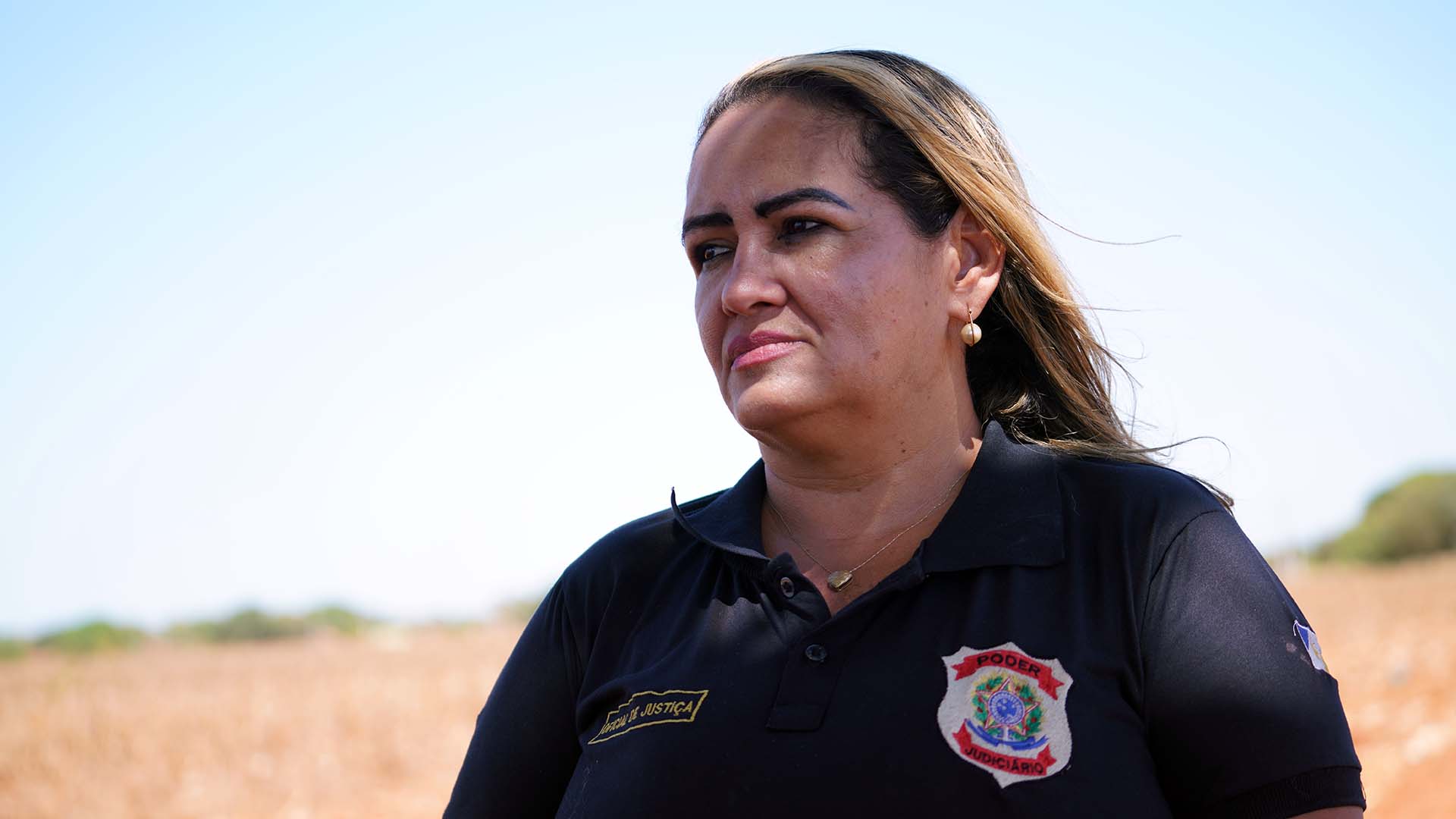 Mulher branca de cabelo loiro e com uma blusa preta; ao fundo uma estrada de terra