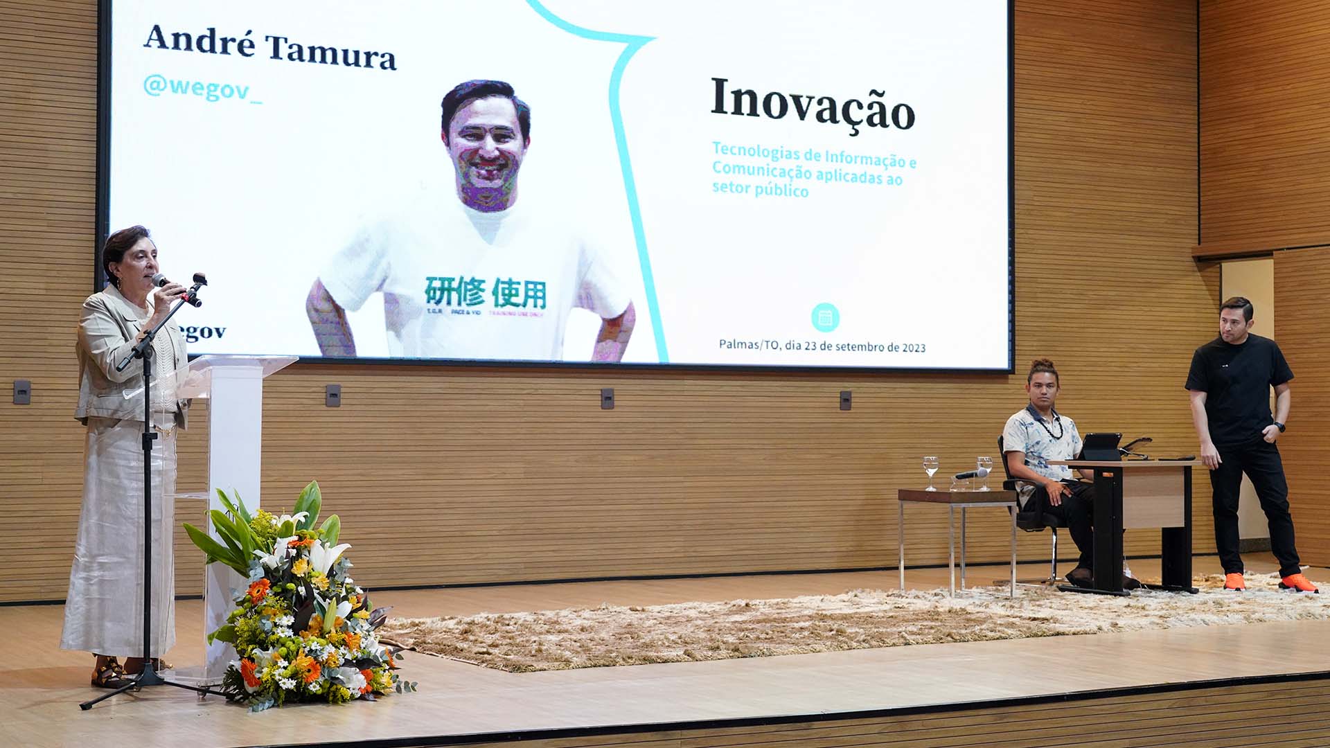 A desembargadora Ângela Haonat fala ao microfone, ela é assistida pela plateia e pelo palestrante que está no palco