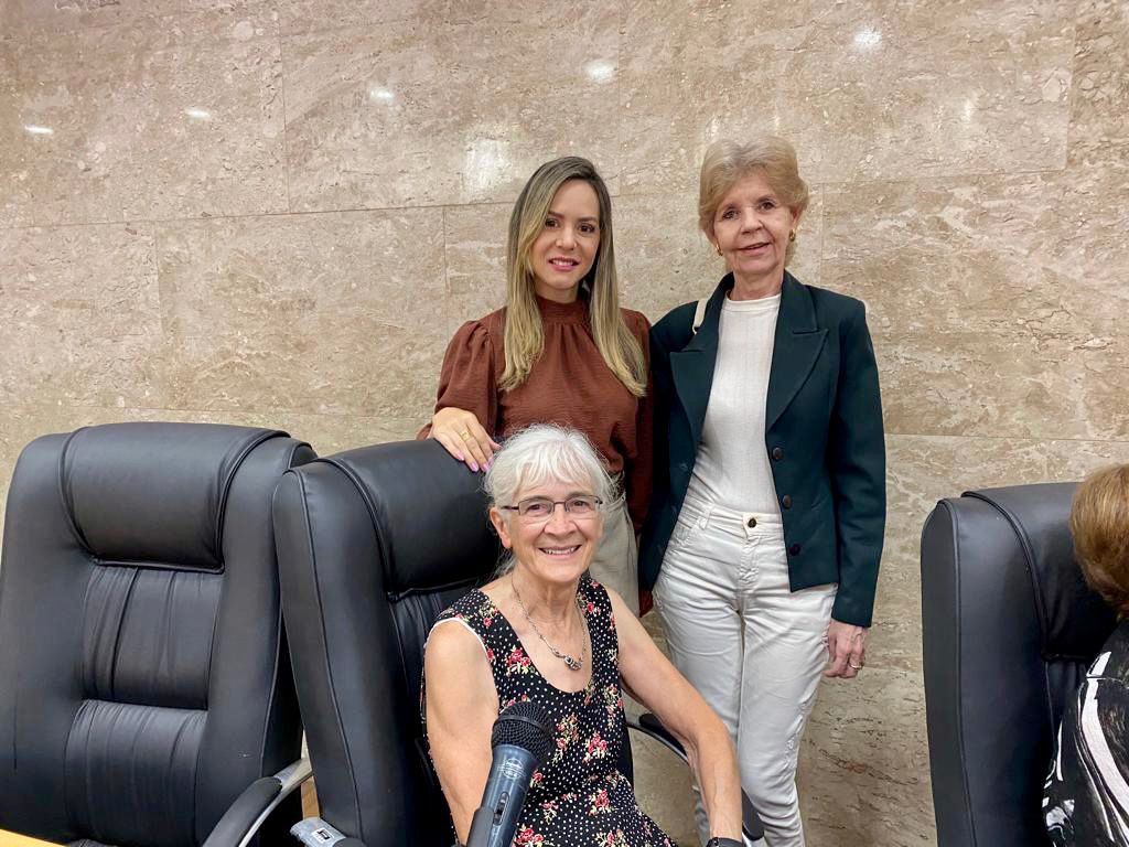 Imagem colorida que mostra duas mulheres loiras em pé e uma senhora grisalha sentada.