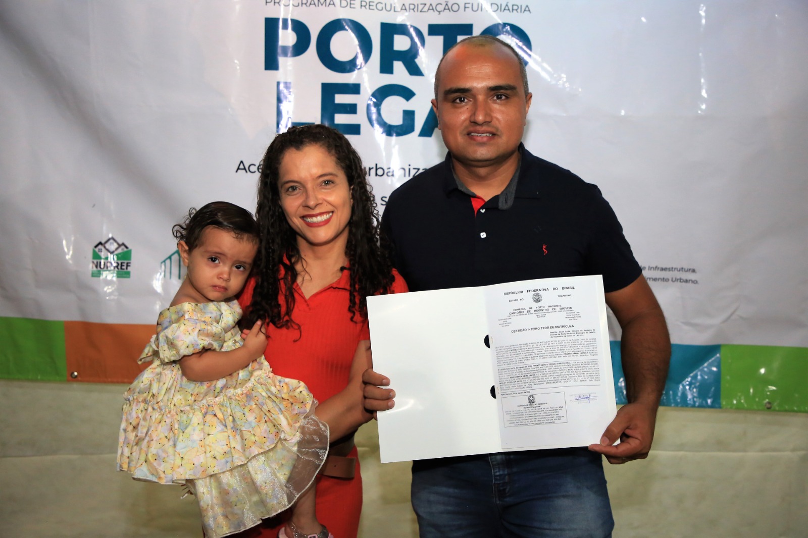 Imagem colorida de um casal posando para foto: a mulher segura uma menina de vestido e o homem segura um documento de regulariação fundiária 