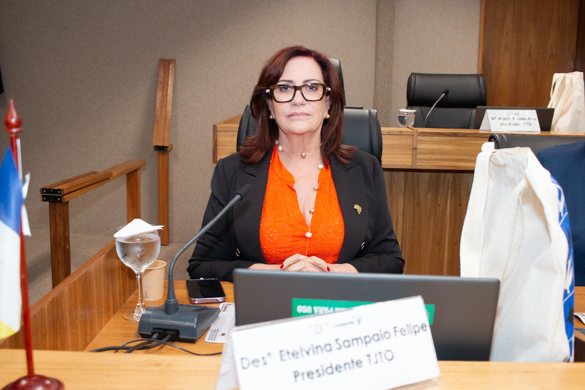 Fotografia colorida que mostra a presidente do TJTO sentada à mesa do Consepre. Ela é uma mulhere branca usando óculos  e camisa laranja e terno preto