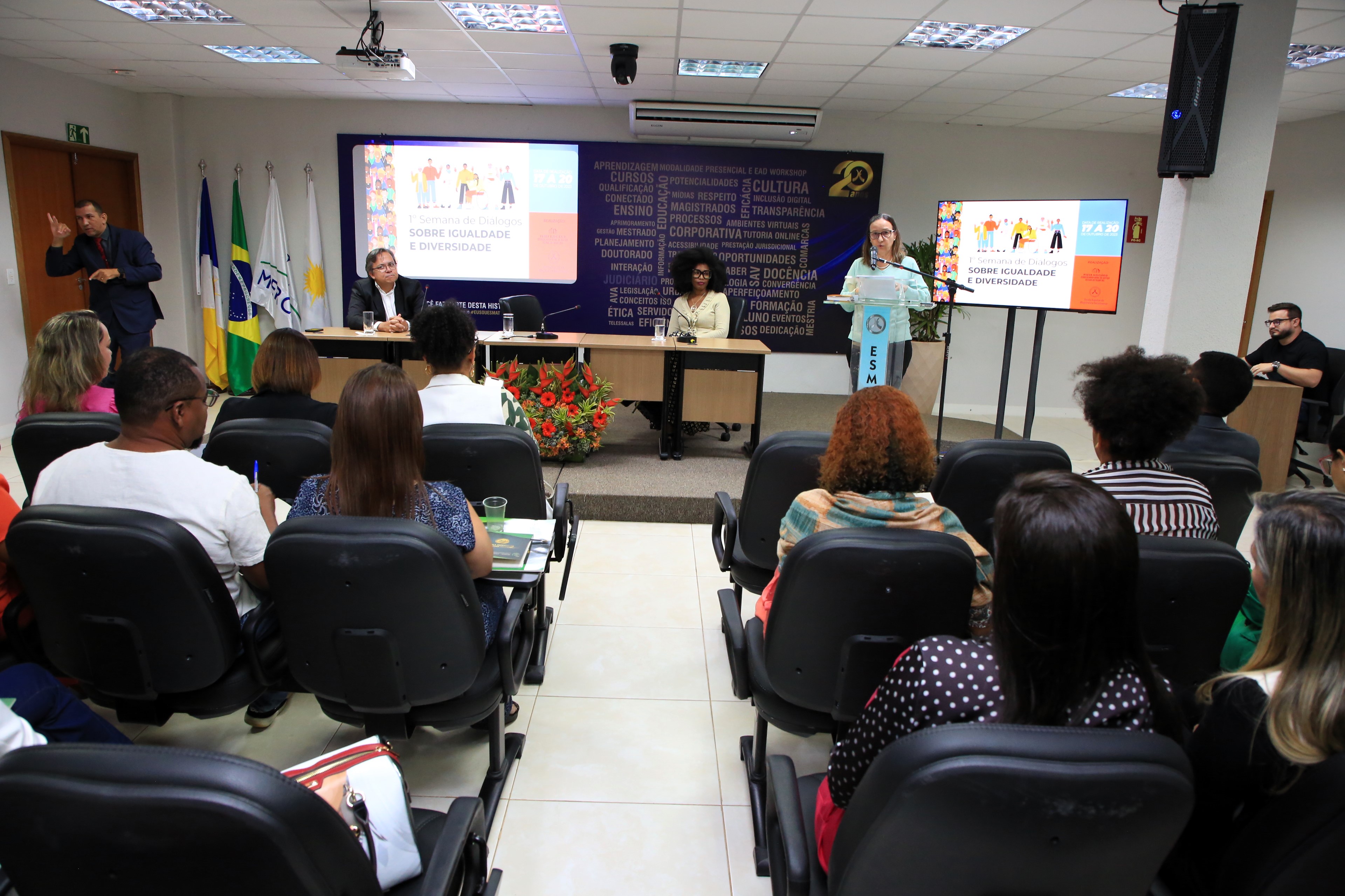 Pessoas sentadas em auditório assistindo palestra de mulher branca de roupa azul, ao lado sentada a mesa, mulher negra de camisa clara e homem pardo de terno escuro