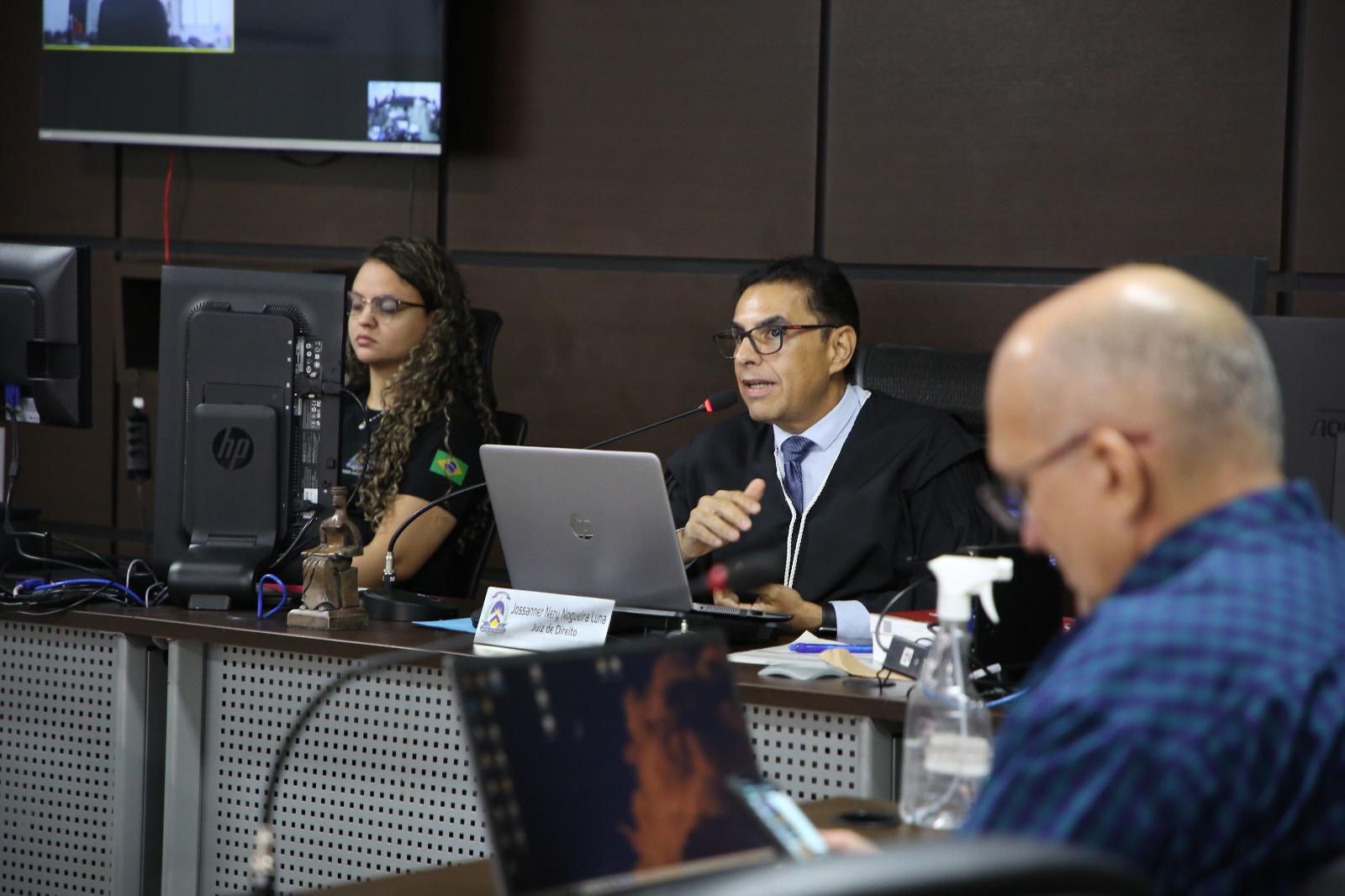 Fotografia colorida de juiz falando ao microfone observados por um homem e uma mulher, dentro do júri 