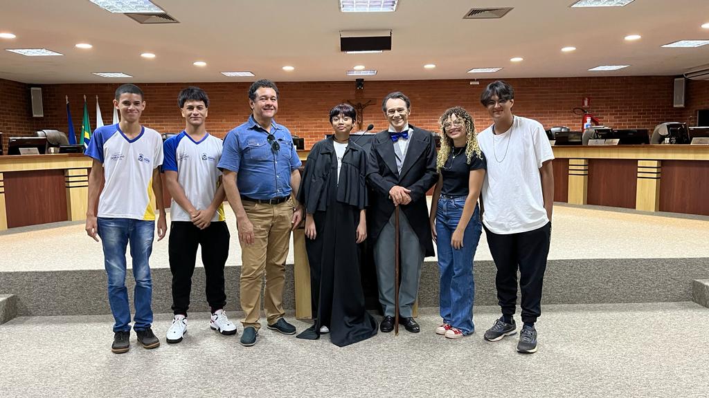 no tribunal do pleno personagem Teotônio veste fraque com camisa branca e gravata azul é acompanhado por estudantes