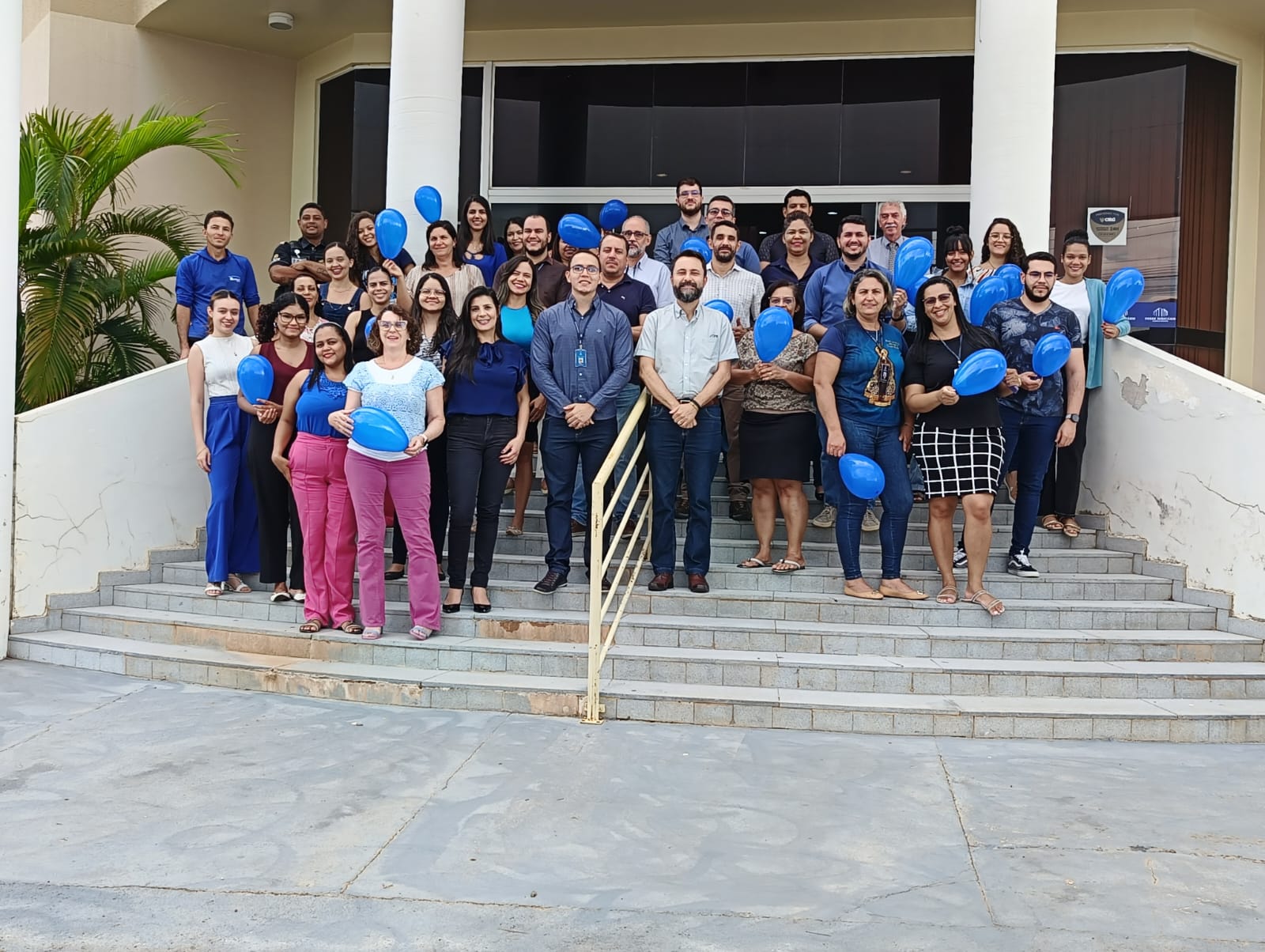 Fotografia colorida de um grupo de pessoas, a maioria vestidos azul e segurando balões azuis, celebrando o apoio da comarca de Dianópolis a  causa do novembro azul nas escadas do fórum da Comarca de Dianópolis