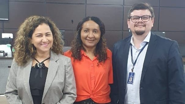 Mulher parda de camisa preta e blazer cinza e cabelos soltos ao lado de mulher  negra, de camisa laranja e cabelo solto ao centro, homem pardo de camisa clara e paletó azul de óculos, cabelos curtos 