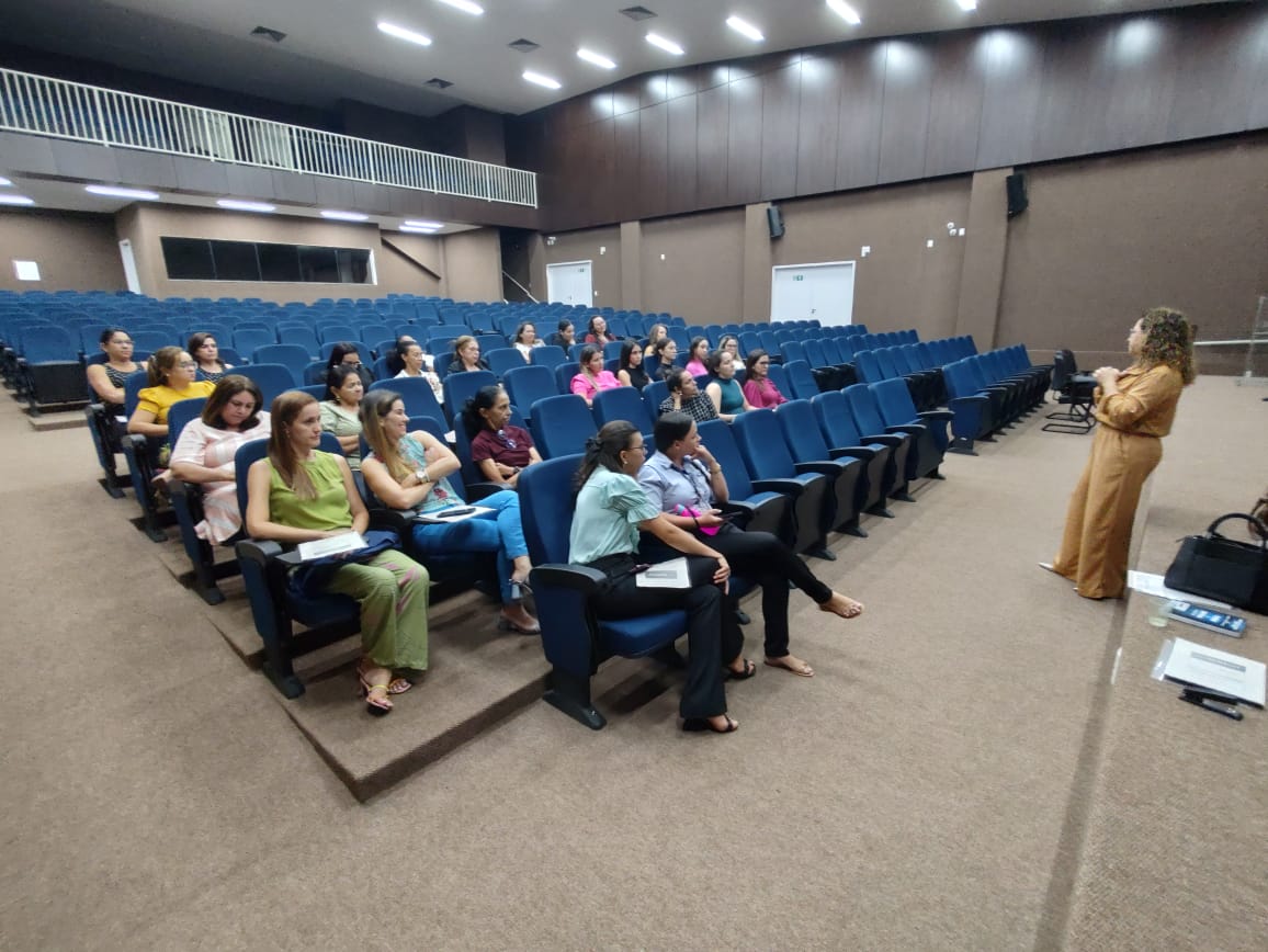 Fotografia colorida onde uma mulher fala, em pé, para um grupo de mulheres sentadas em um auditório 