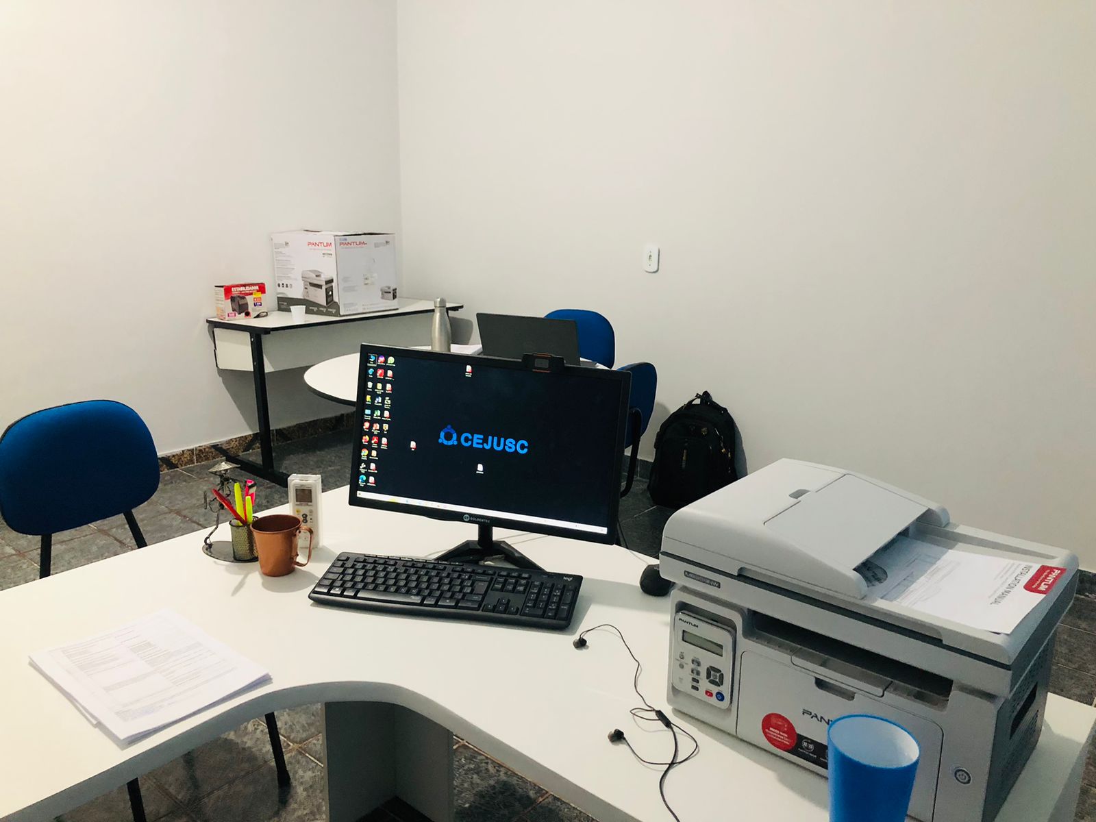 Imagem mostra uma mesa clara, com um computador e uma impressora; existe também uma cadeira azul
