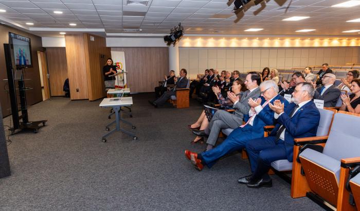 Imagem mostra um auditório com pessoas sentadas em poltronas cinzas e à frente um telão