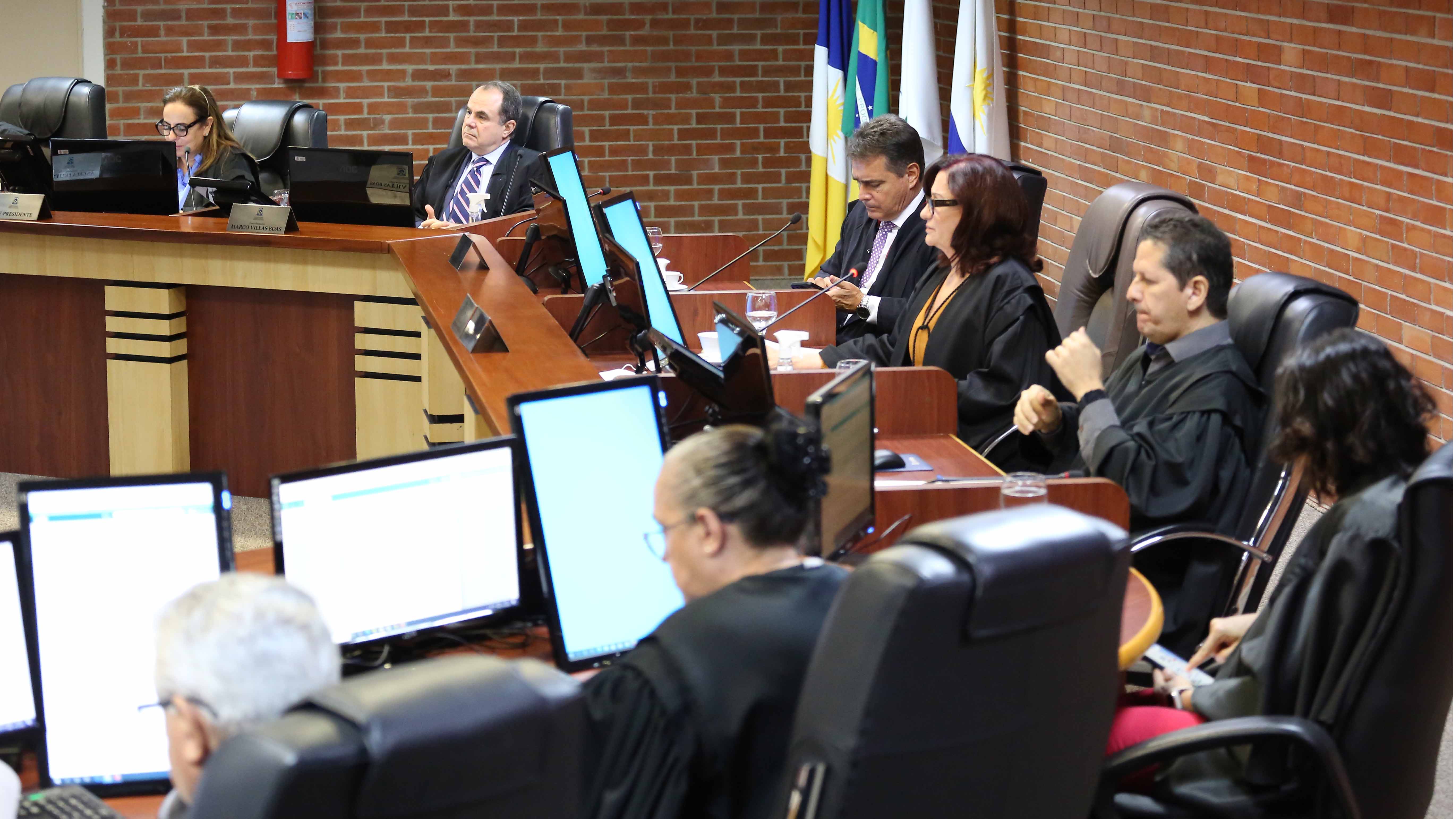 Imagem mostra uma sala com tijolinhos à vista em frente uma mesa redonda e pessoas sentadas com computador  na mesa 