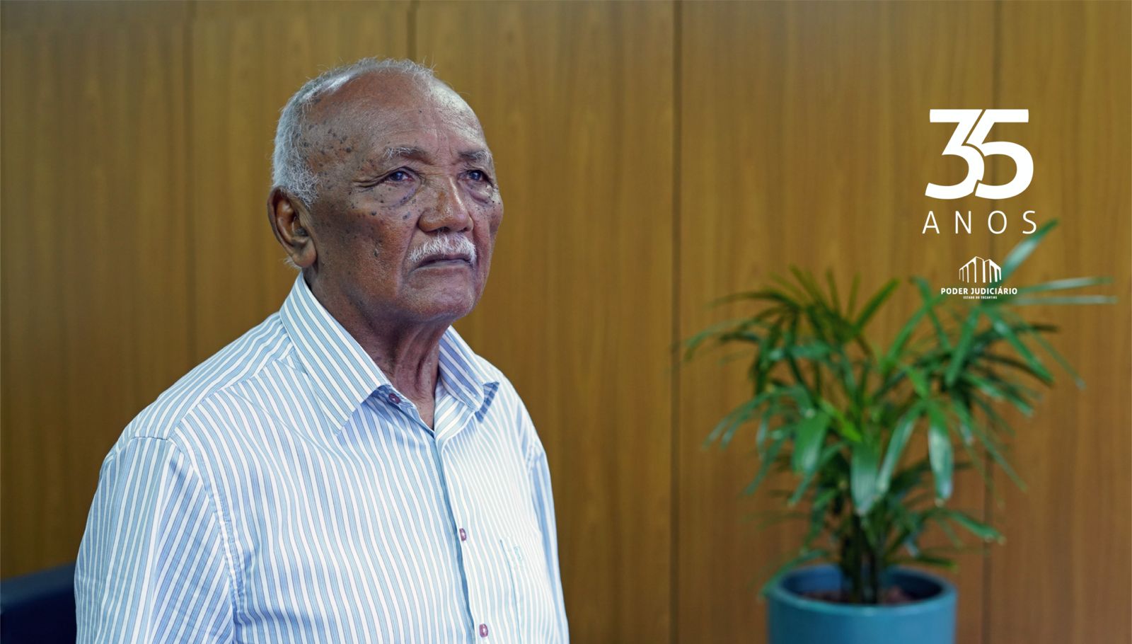 Homem negro, idoso, parcialmente calvo e cabelos e bigode grisalhos, de camisa branca. Ao fundo painél de madeira com vaso de planta ao lado