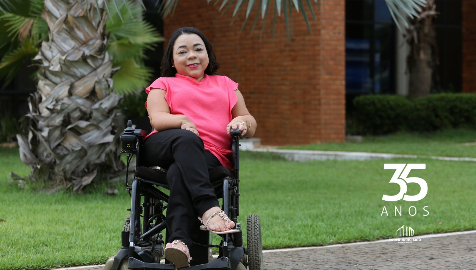 Imagem mostra ao fundo um jardim e à frente uma mulher cadeirante vestida com calça preta e blusa rosa
