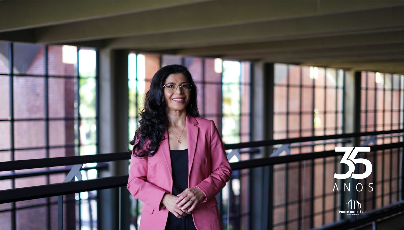 Foto mostra uma mulher com cabelos longos, em pé e com roupa preta e rosa; ao fundo janelas de vidro