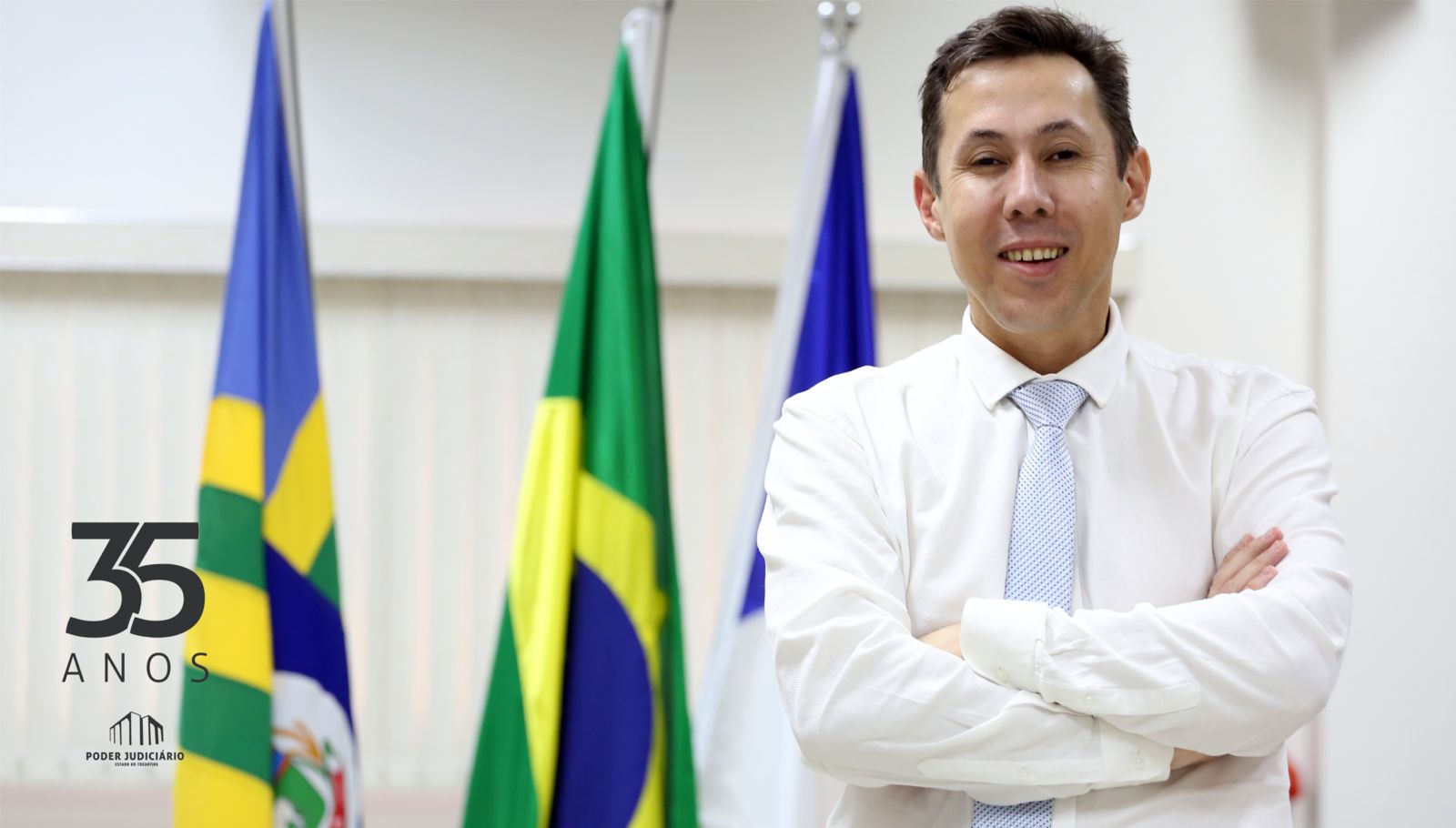 Homem branco de camisa e gravata azul clara, de braços cruzados, ao fundo parede branca e mastro com bandeiras