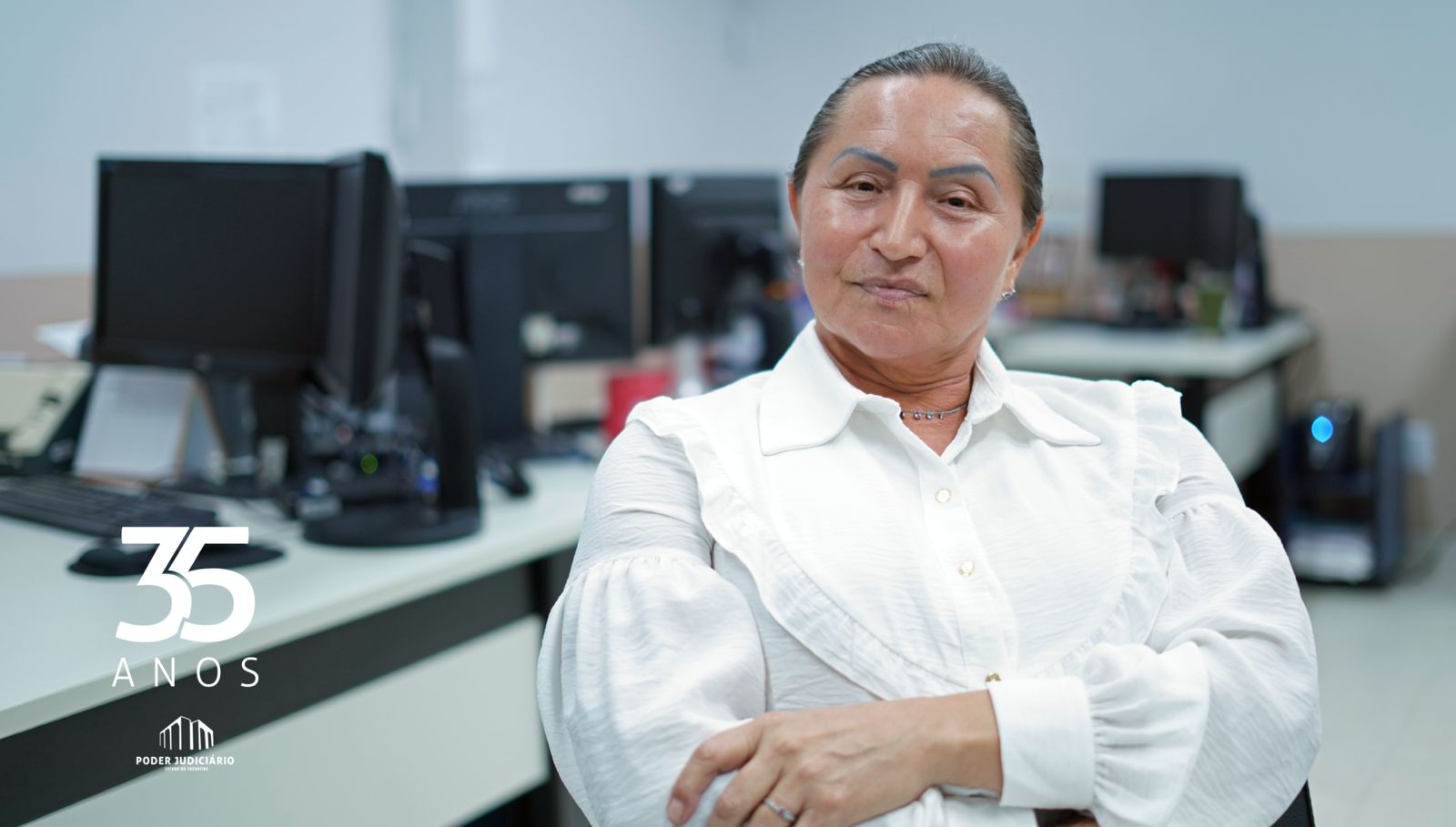 Foto mostra uma mulher com cabelos presos, camisa branca, braços cruzados e sentada em uma cadeira; ao fundo computadores