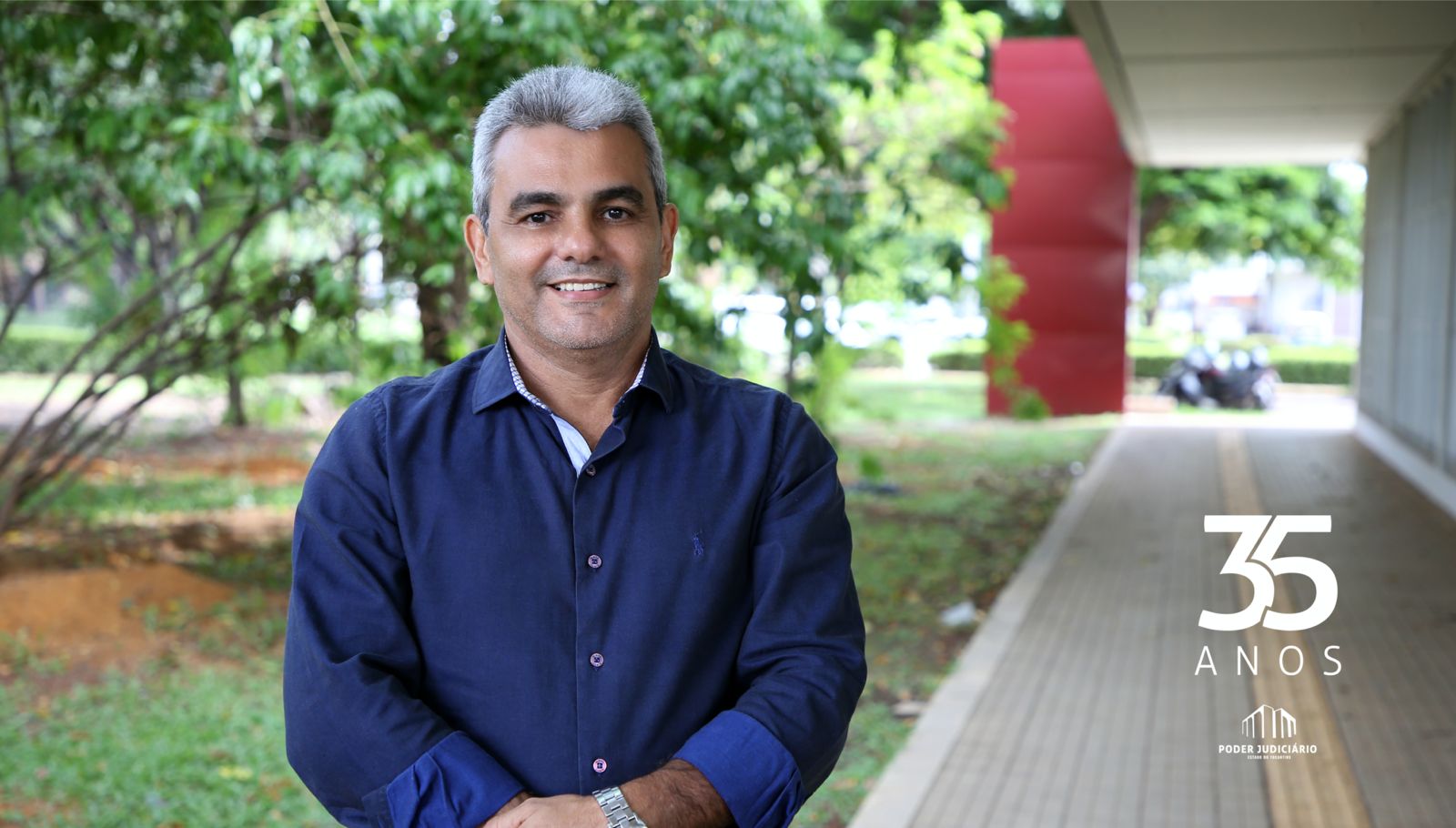 Imagem mostra um homem de cabelo branco, blusa azul e braço cruzado; ao fundo paisagem de um jardim