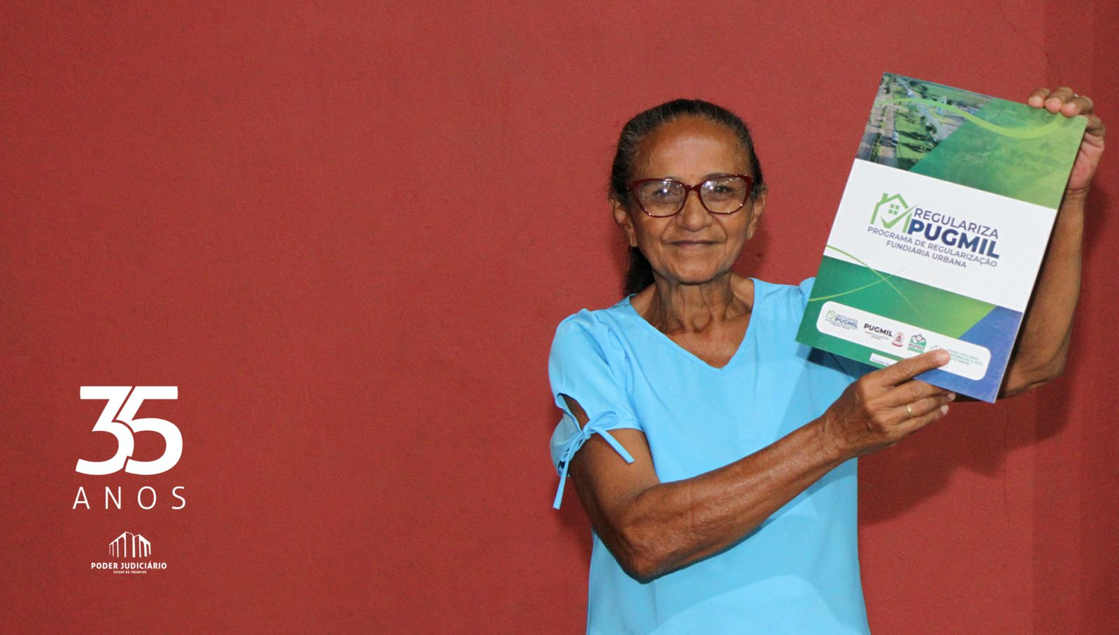 Mulher idosa de cabelo amarrado vestida de camisa azul clara de manga curta, segurando ao alto o título de regularização de sua casa. Ela está encostada numa parede vermelha.