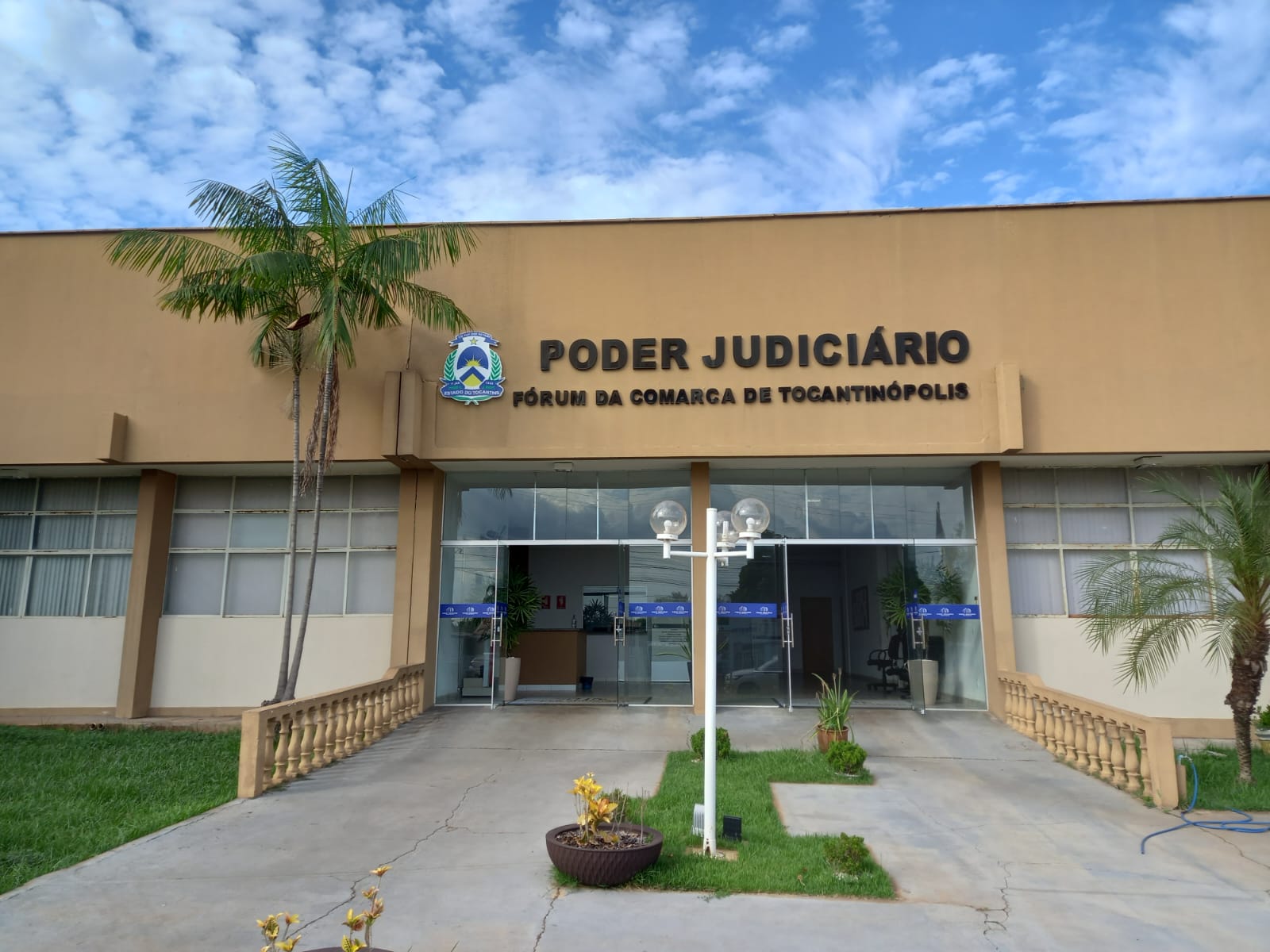 Fachada do prédio do Fórum de Tocantinópolis na cor bege com palmeira na frente. Céu azul com pequenas nuvens brancas