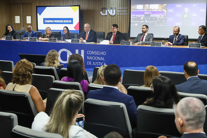 Fotografia colorida da Cerimonia de entrega do 3º Prêmio Prioridade Absoluta, onde pessoas estão sentadas em cadeiras na plateia enquanto os ministrantes da premiações estão em uma mesa longa em sua frente